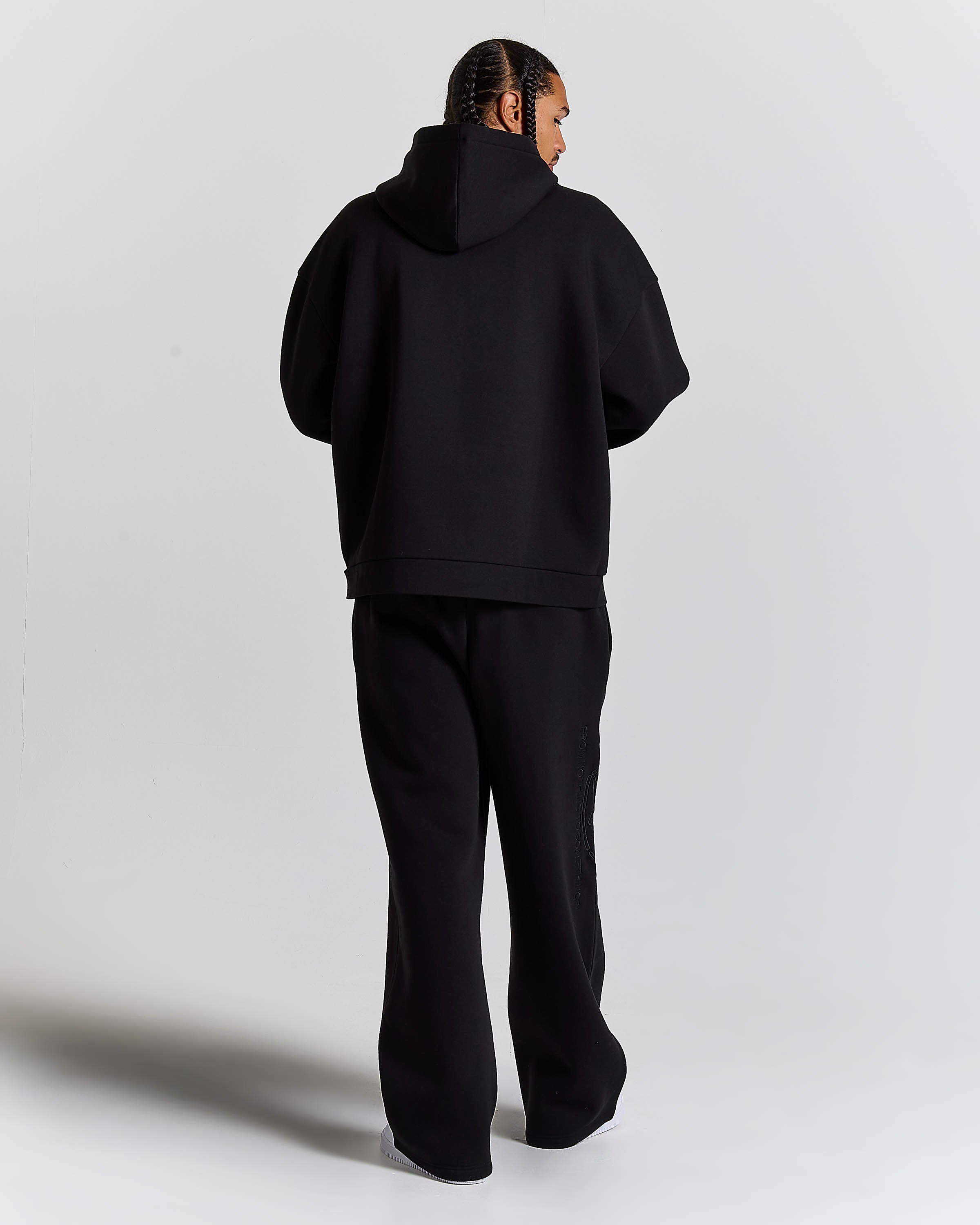 A model viewed from behind wearing a black oversized hoodie and matching black wide-leg sweatpants against a light gray studio background.