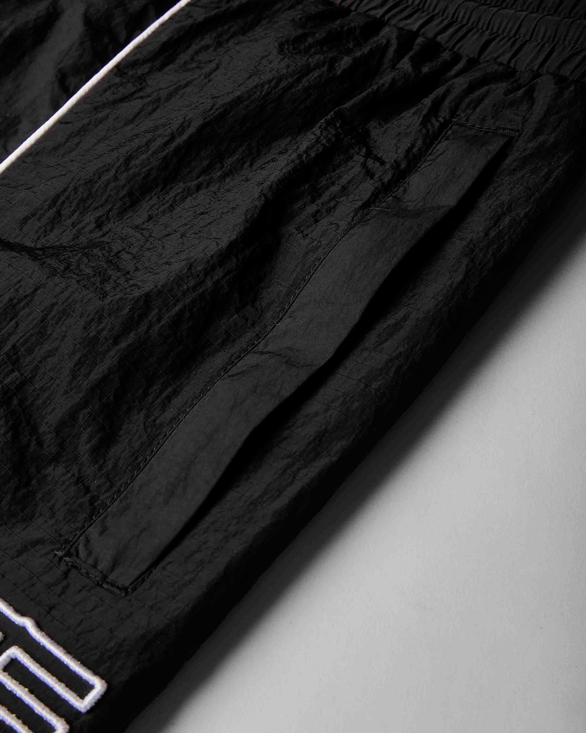 Close-up of black crinkled lightweight fabric with a white outlined logo at the bottom left, photographed on a light gray background.