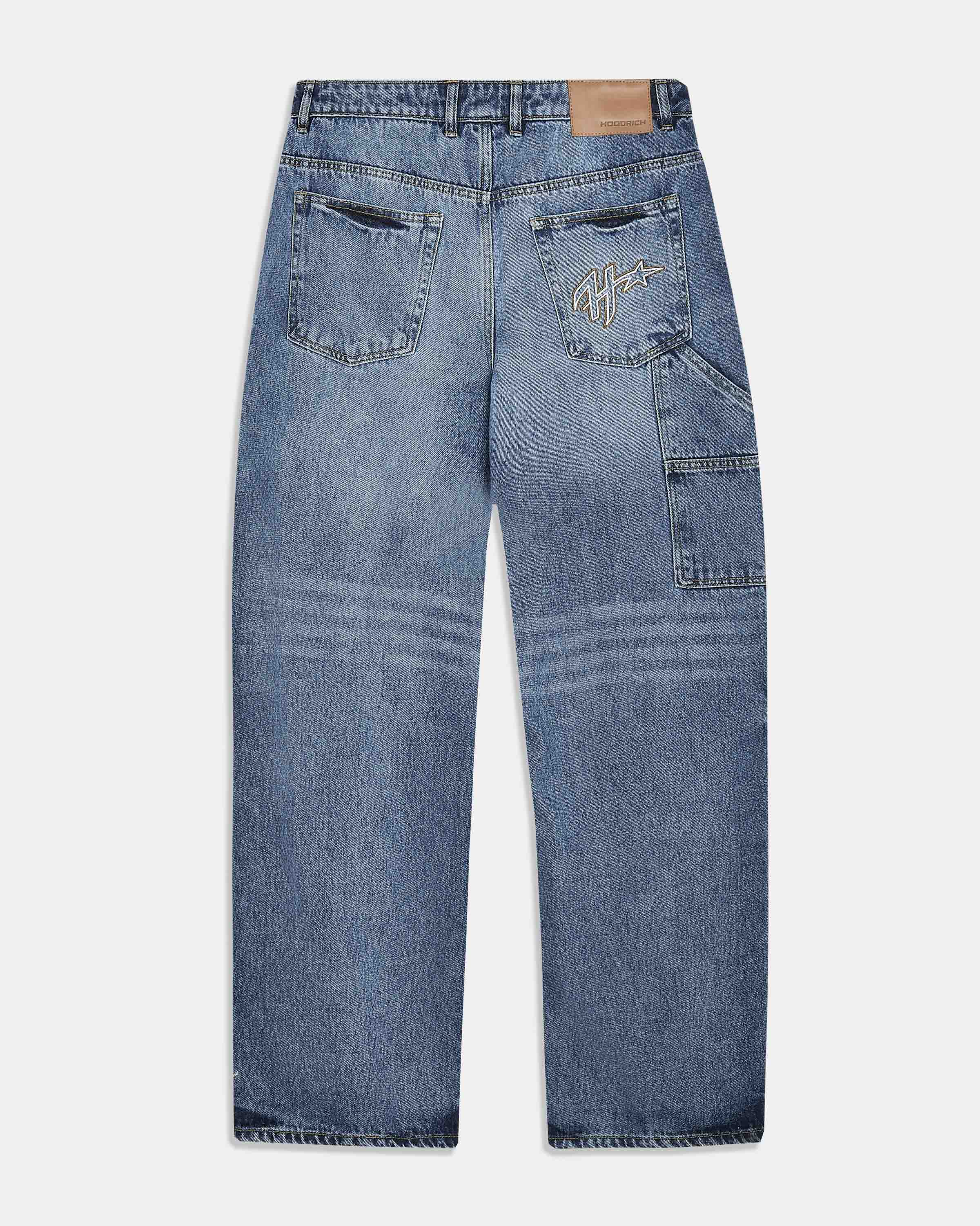 Back view of a pair of blue denim jeans with a light wash, featuring a tan leather patch on the back waistband and a stylized embroidered logo on the right back pocket.