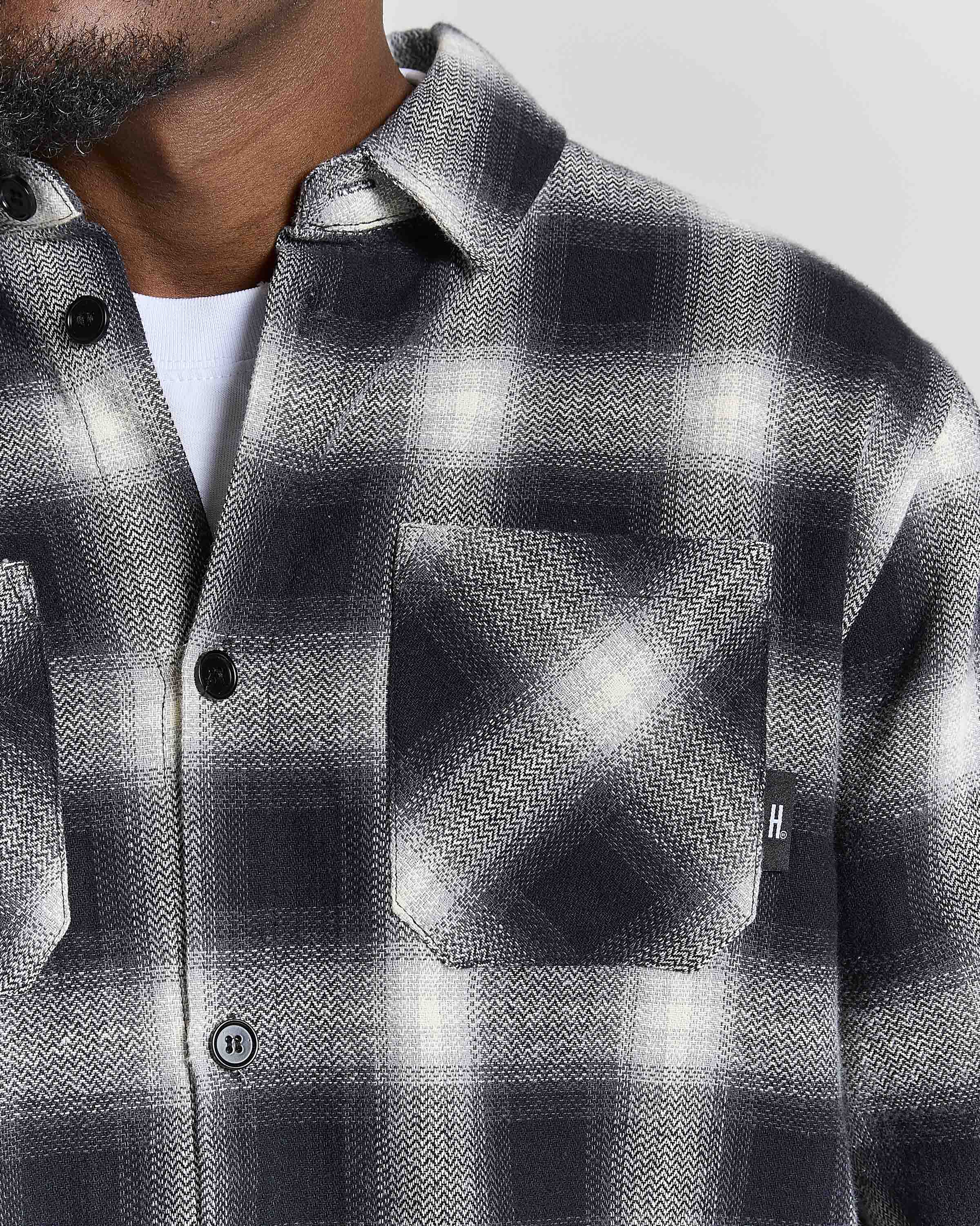 Close-up of a man wearing a black, white, and gray plaid button-up shirt with a chest pocket, layered over a white undershirt.