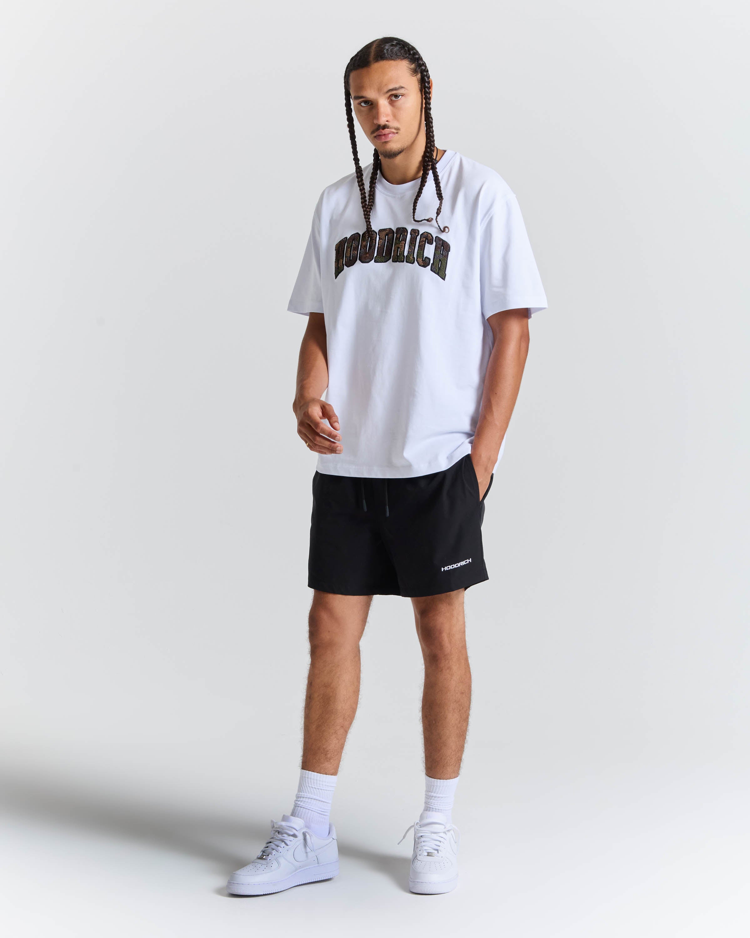 A man modeling a white short-sleeve t-shirt with 'HOODRICH' printed in black across the chest, paired with black athletic shorts, white socks, and white sneakers, against a light gray studio background.