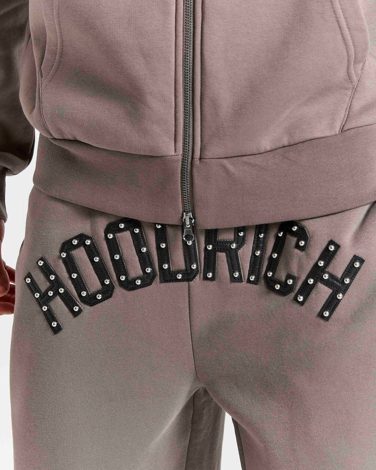 Displays a model wearing the Brown 'Hoodrich' branded men's Varso Studs Full Tracksuit featuring the details on the hoodrich branding