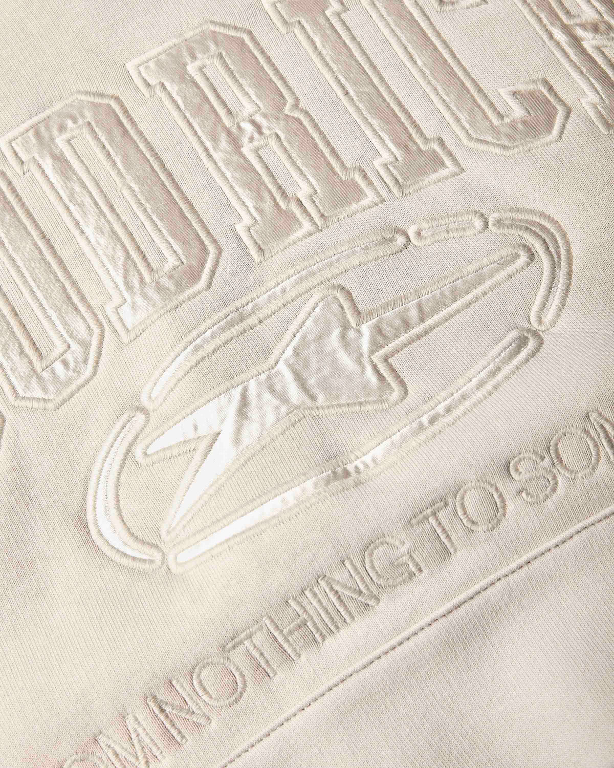 Close-up of a cream-colored fabric surface with tonal, raised embroidered lettering and a lightning bolt graphic.