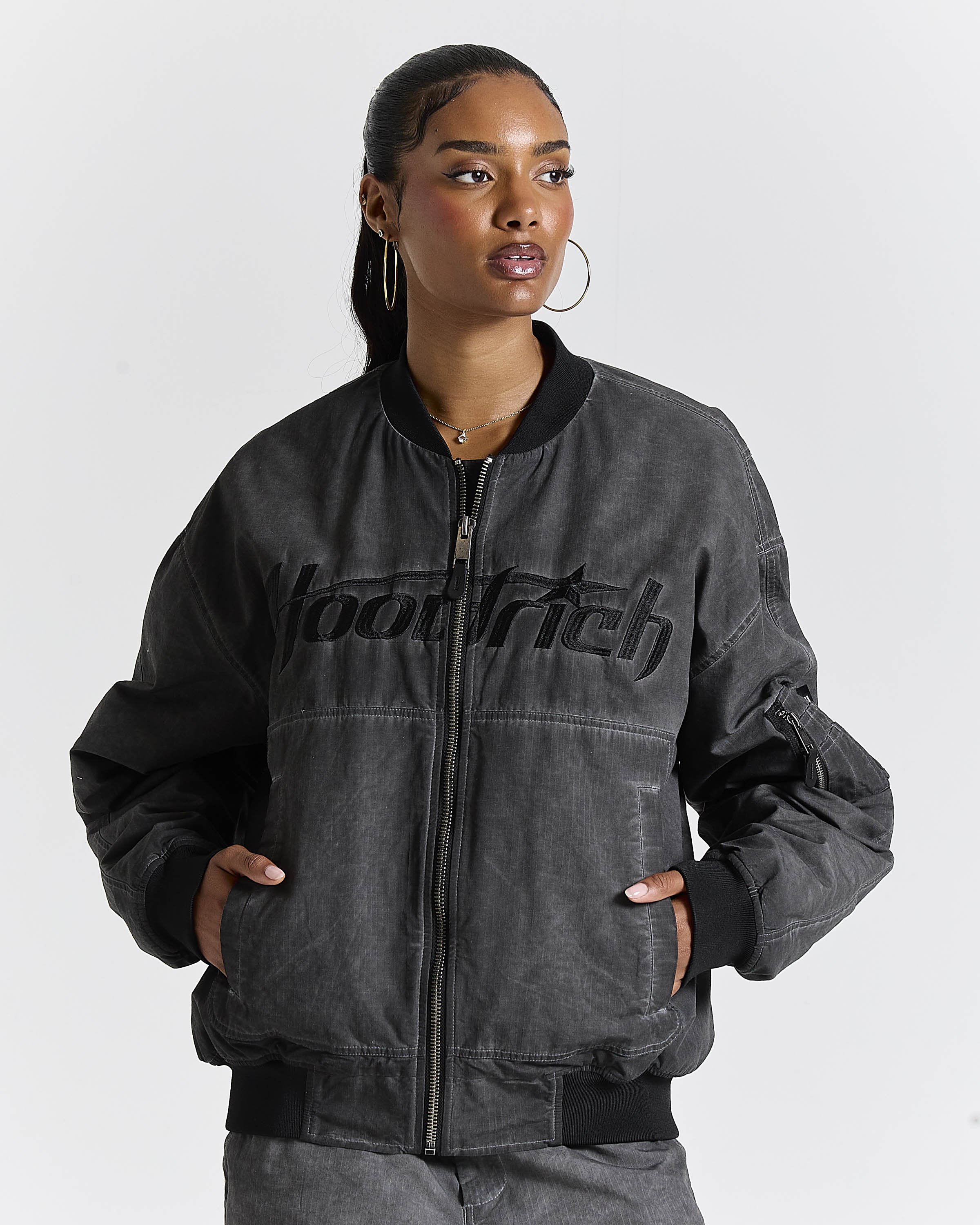 A woman wearing a dark gray washed bomber jacket with black ribbed trim and a black 'hoodrich' logo across the chest, photographed against a light gray studio background.