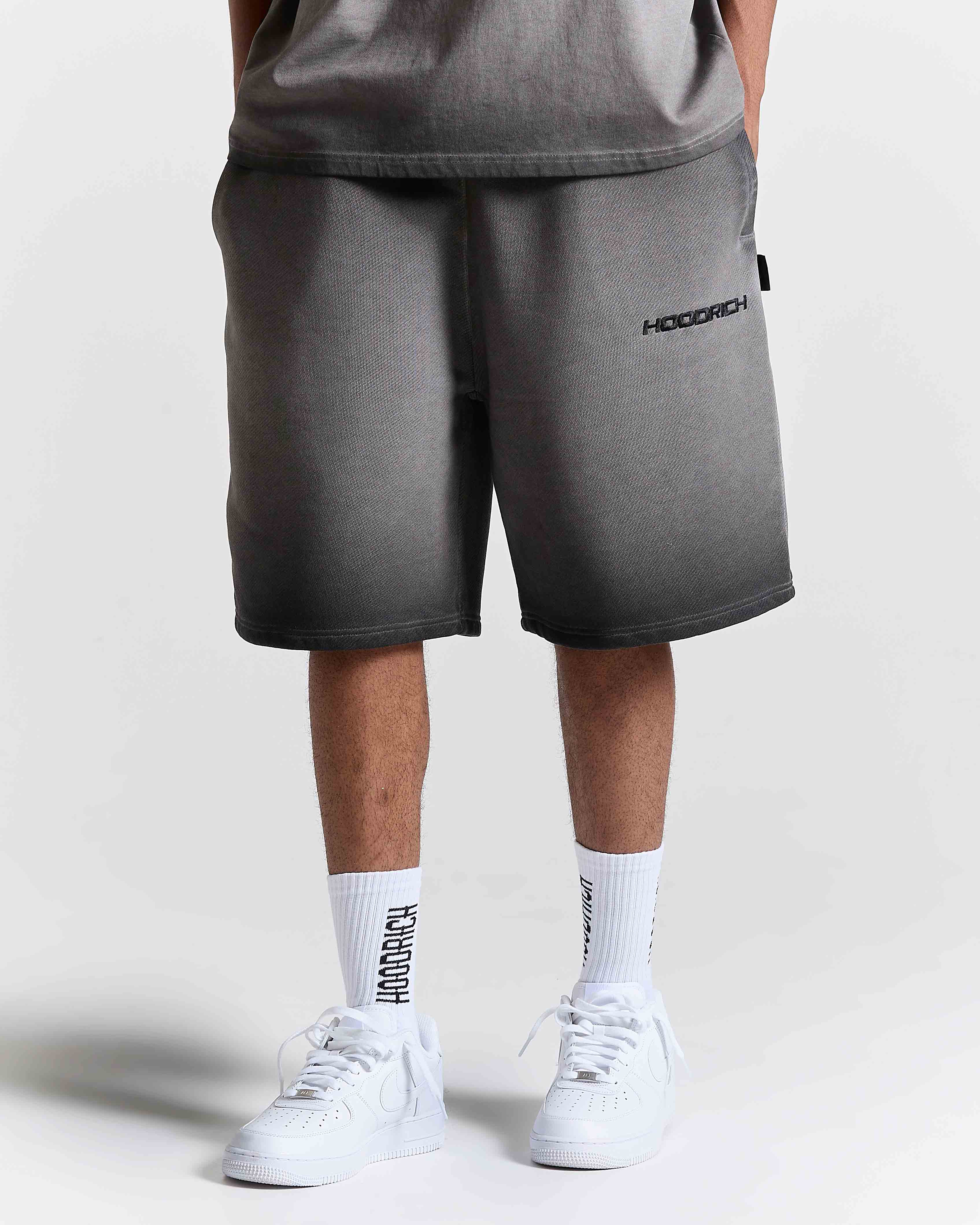 Model wearing oversized black oil wash shorts with Hoodrich logo embroidery, white socks, and white trainers.