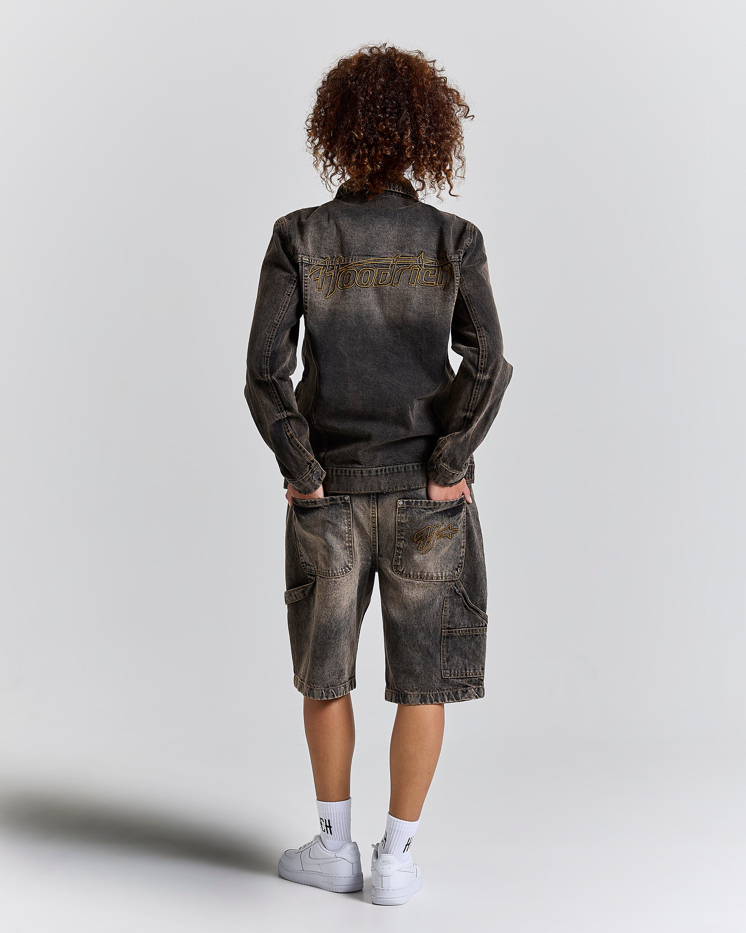 A model viewed from behind wearing a washed black denim jacket and matching distressed denim shorts, paired with white socks and sneakers, against a light gray studio background.