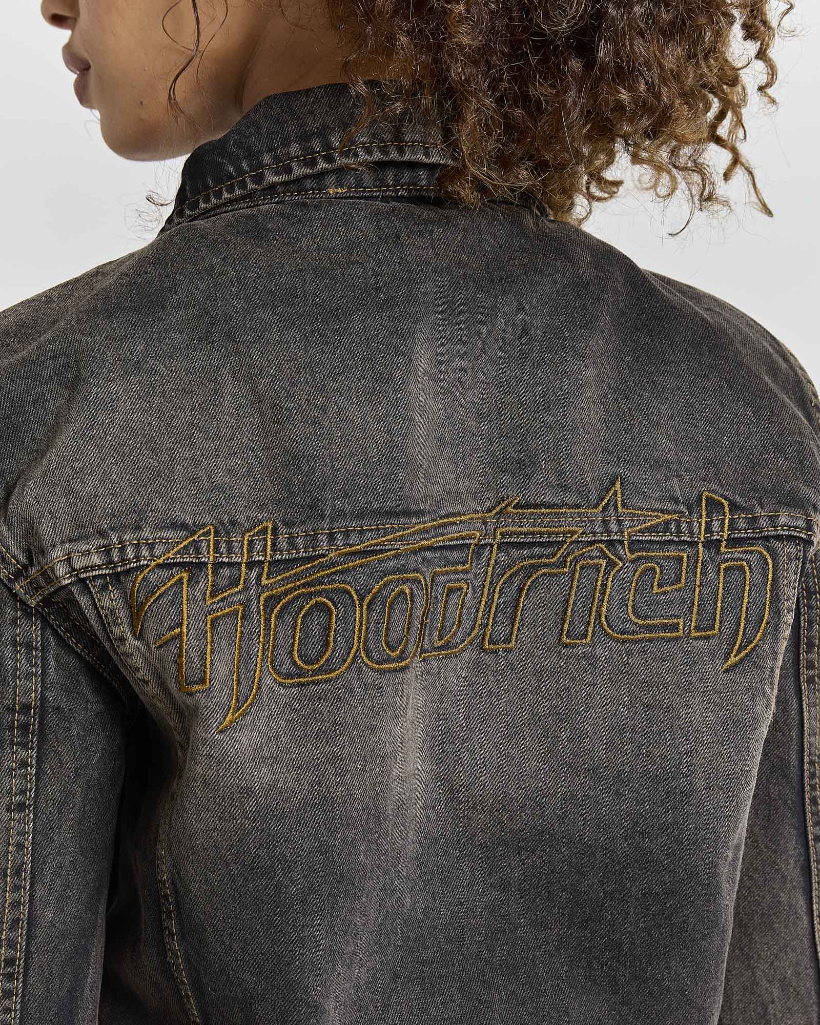 Close-up back view of a person wearing a dark gray washed denim jacket with a large metallic gold embroidered 'Hoodrich' logo across the upper back.