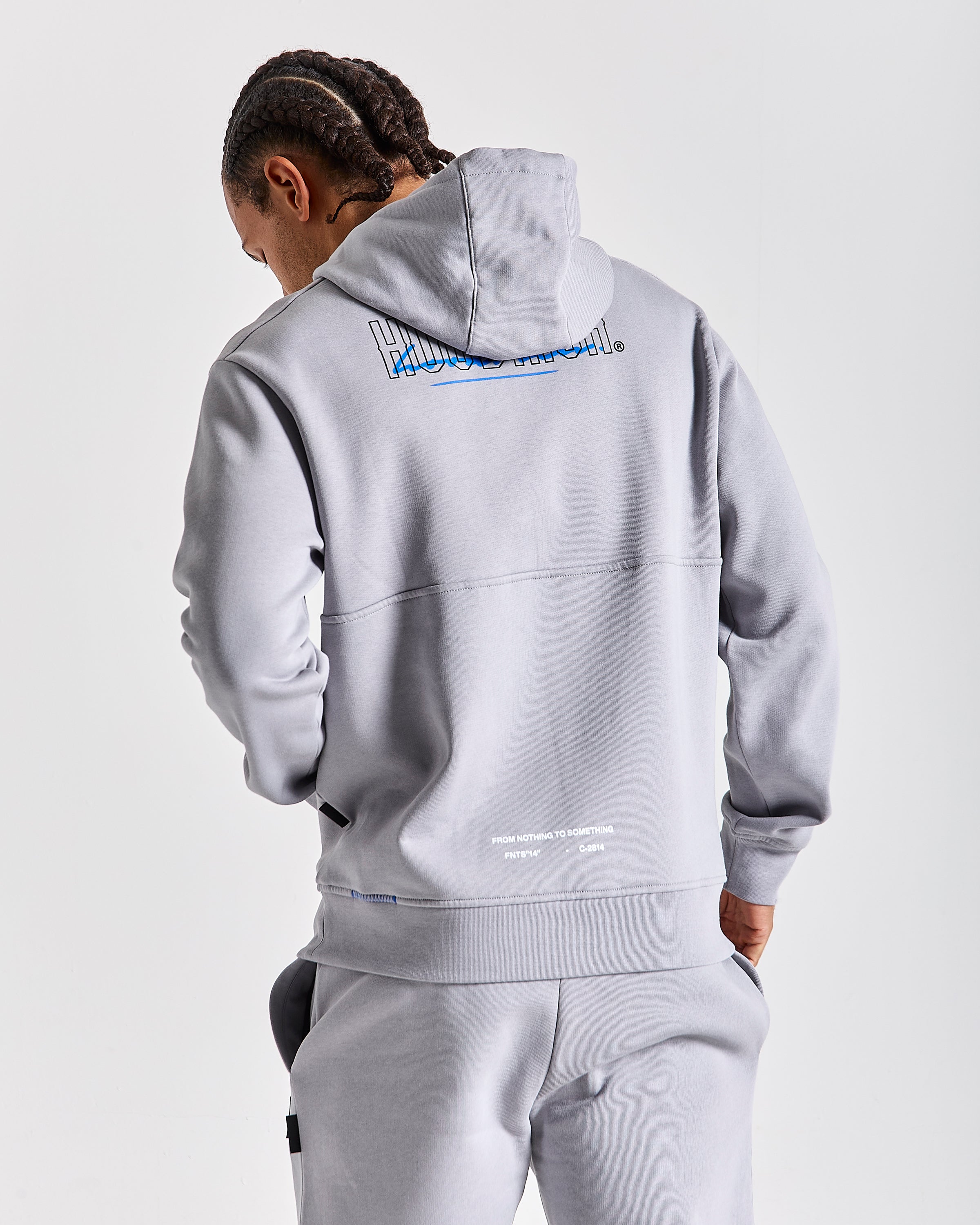 A man wearing a light gray pullover hoodie and matching sweatpants, shown from the back against a light gray studio background. The hoodie features a large blue and white graphic on the upper back and small white text near the lower back.