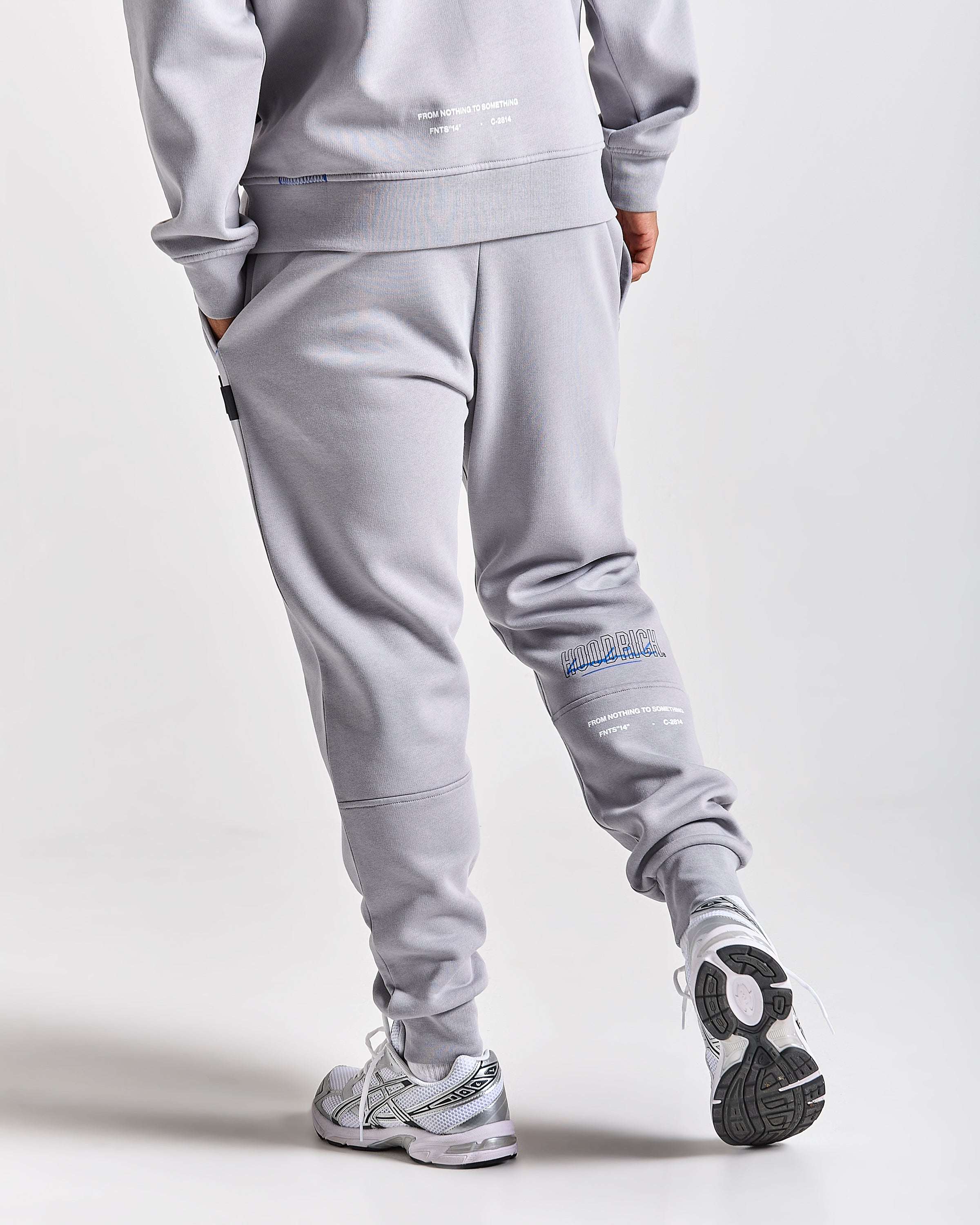 A person wearing light gray sweatpants and a matching sweatshirt, shown from the back in a studio setting. The sweatpants have small white and blue graphic text details near the lower leg, and the person is wearing white and gray sneakers.