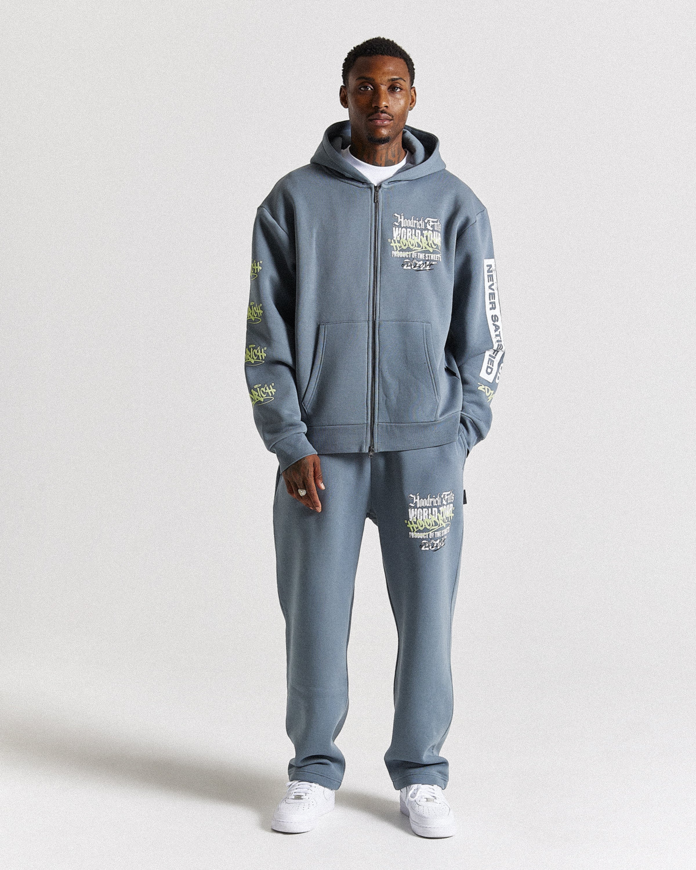 A man modeling a matching slate blue zip-up hoodie and sweatpants set with white sneakers, photographed against a neutral light gray studio background.