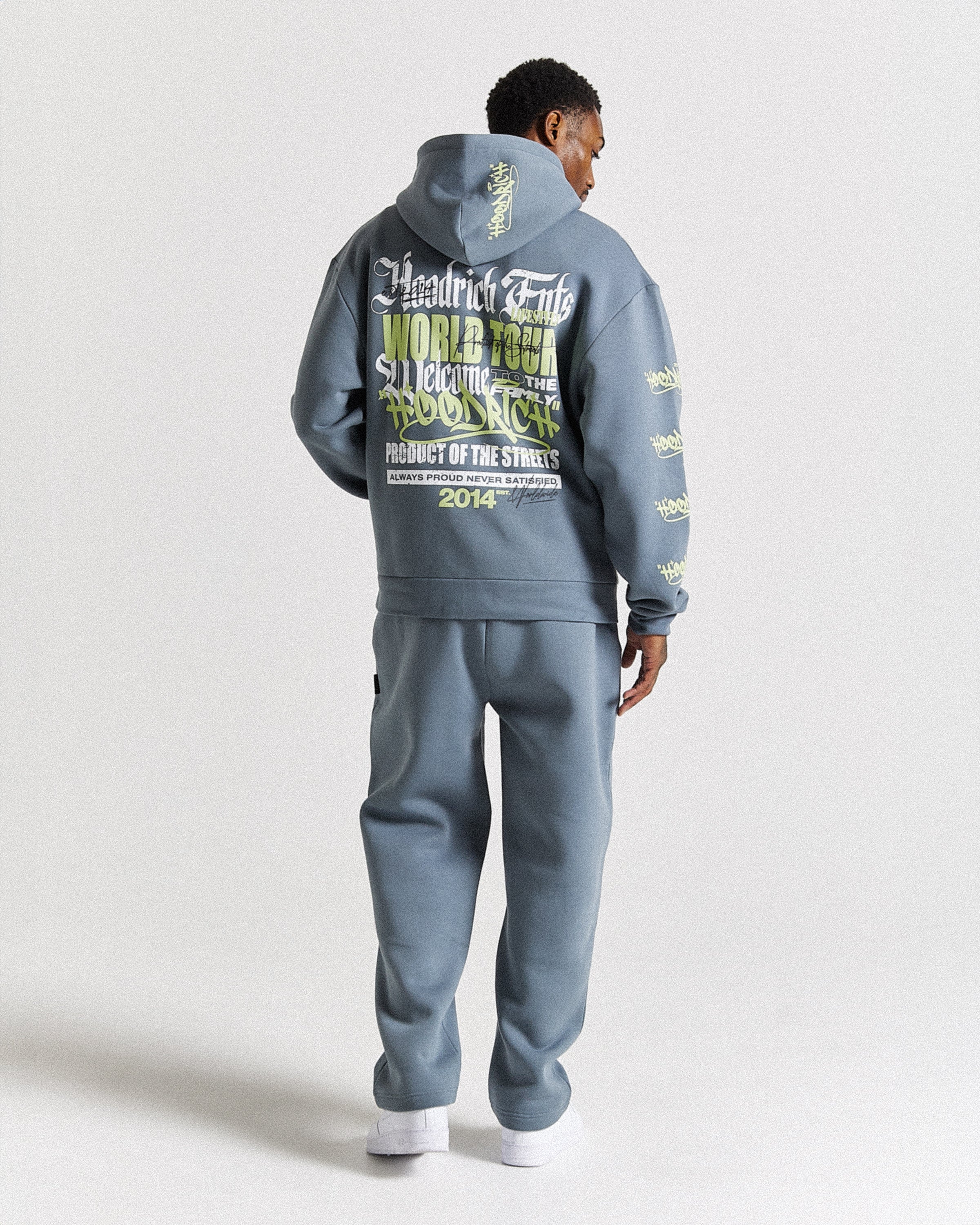 A man viewed from behind wearing a slate blue-gray tracksuit with a large graphic print on the back and smaller prints on the sleeves, paired with white sneakers against a light gray studio background.