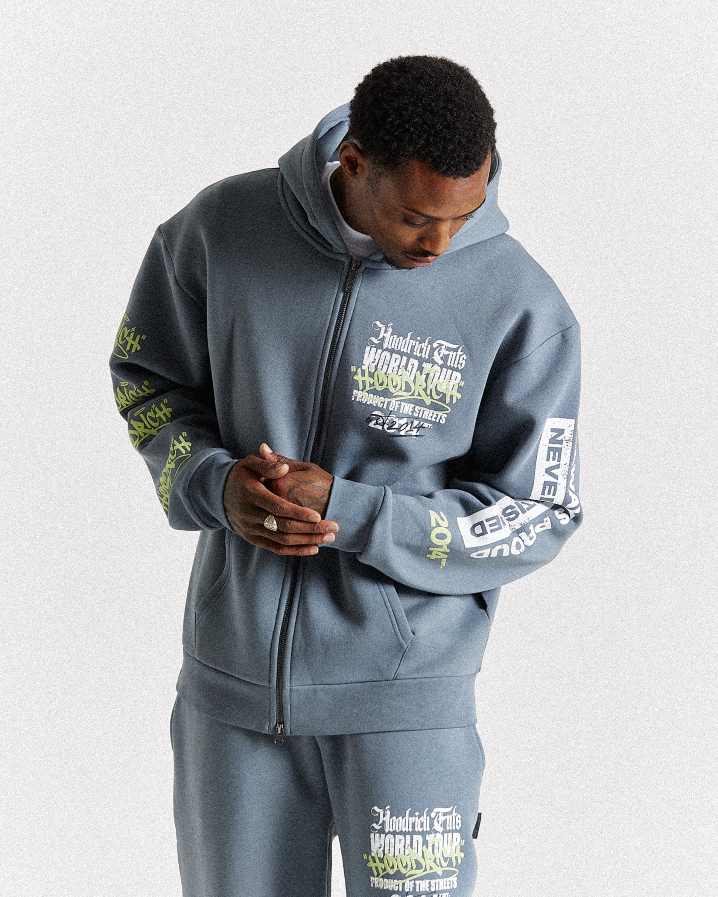 A man wearing a matching gray zip-up hoodie and sweatpants set with white and green graphic text details, photographed against a light gray studio background.