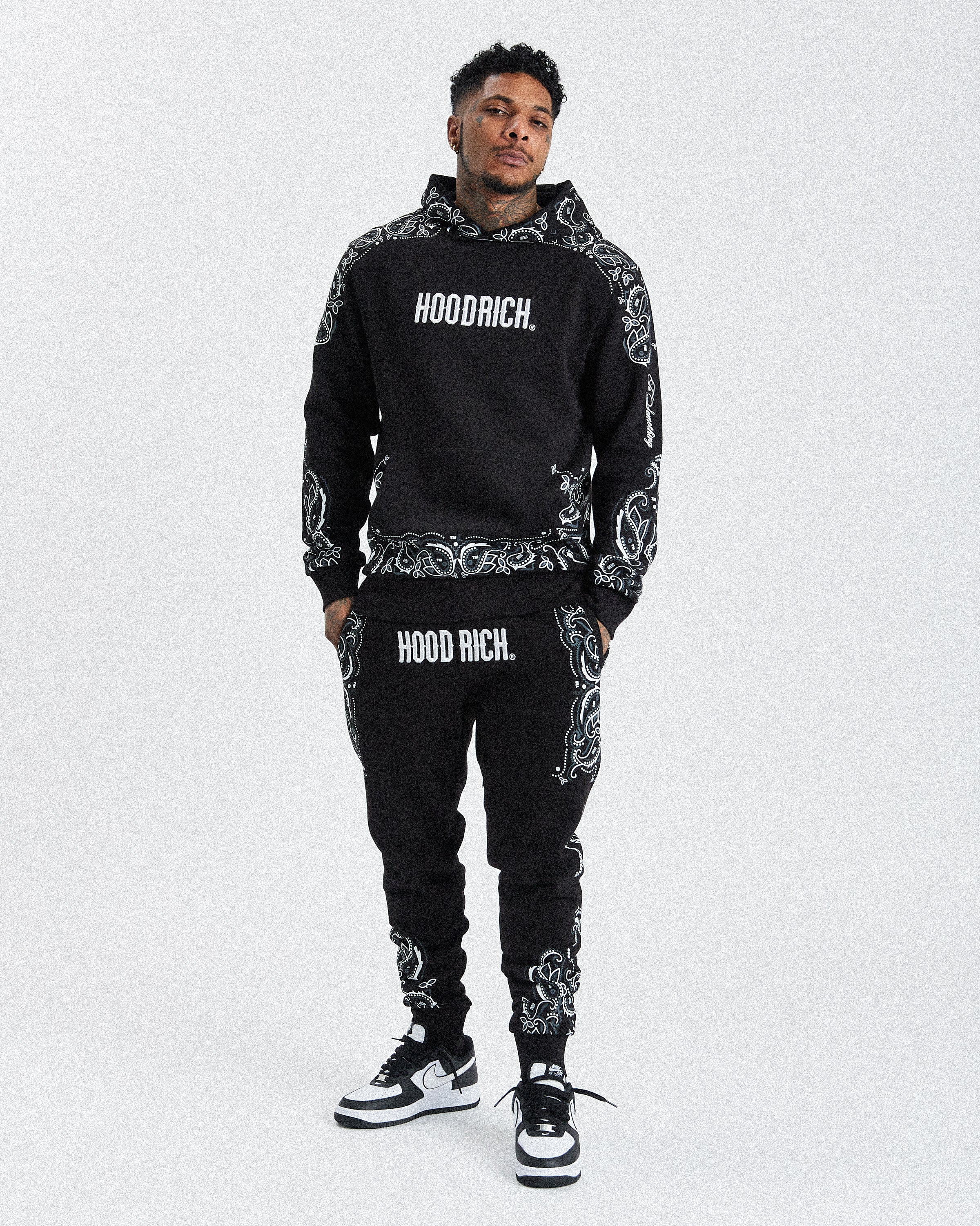A man modeling a black and white patterned hoodie and matching sweatpants with 'HOODRICH.' printed in white on the chest and thighs, paired with black and white sneakers, against a light gray background.