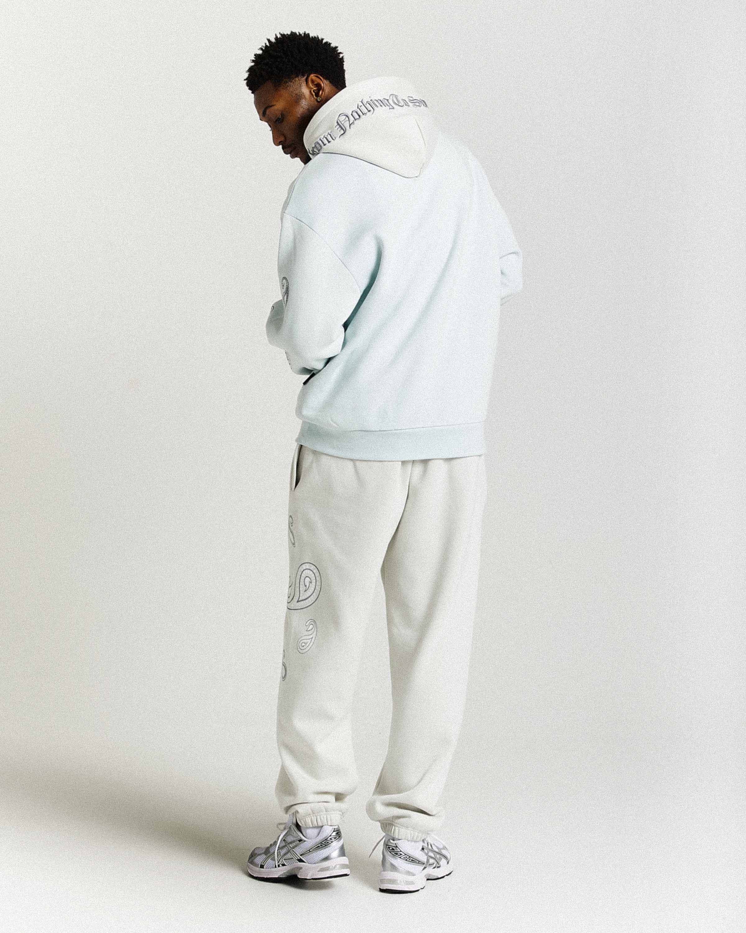 Person wearing a light blue hoodie and white pants with brand logos on a plain background