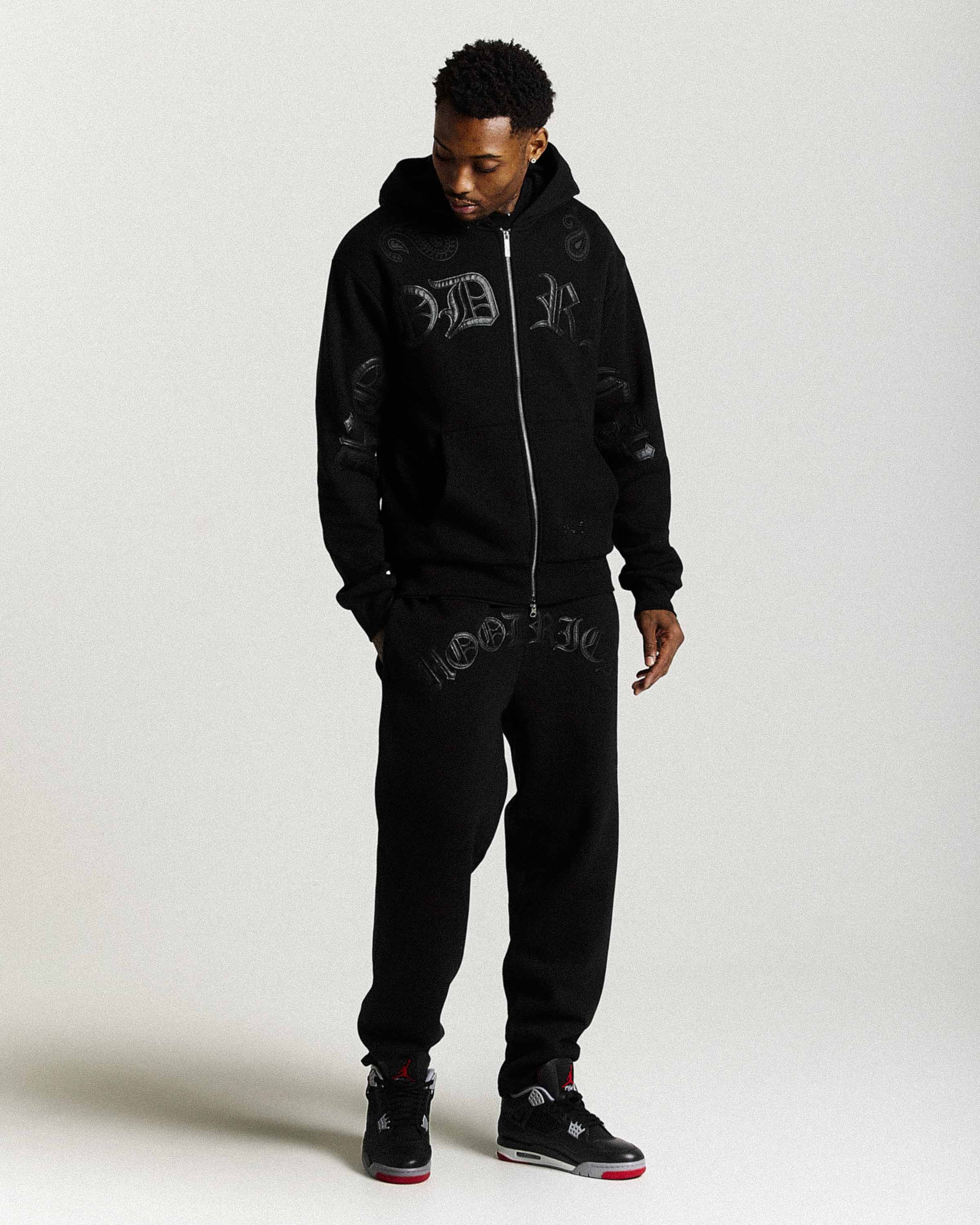 Person wearing a black hoodie and pants with visible branding on a white background