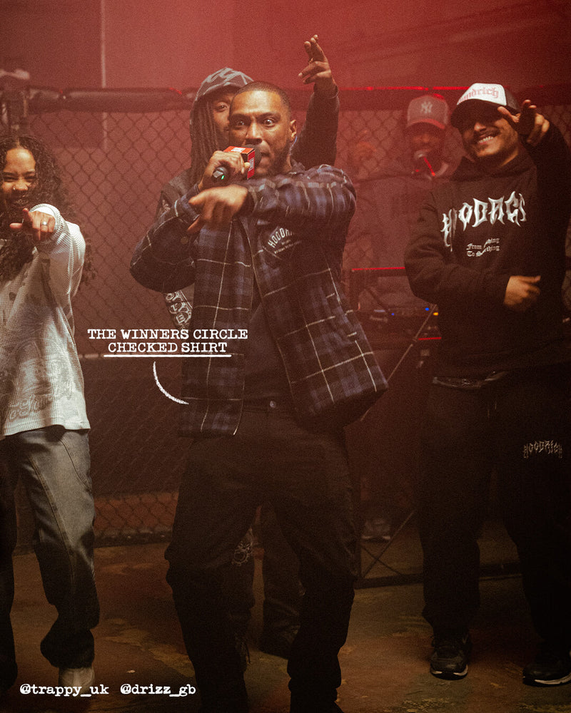 A group of five men posing together in a dimly lit, red-hued urban setting in front of a chain-link fence. The central figure wears a dark plaid flannel shirt and black pants, holding a microphone.