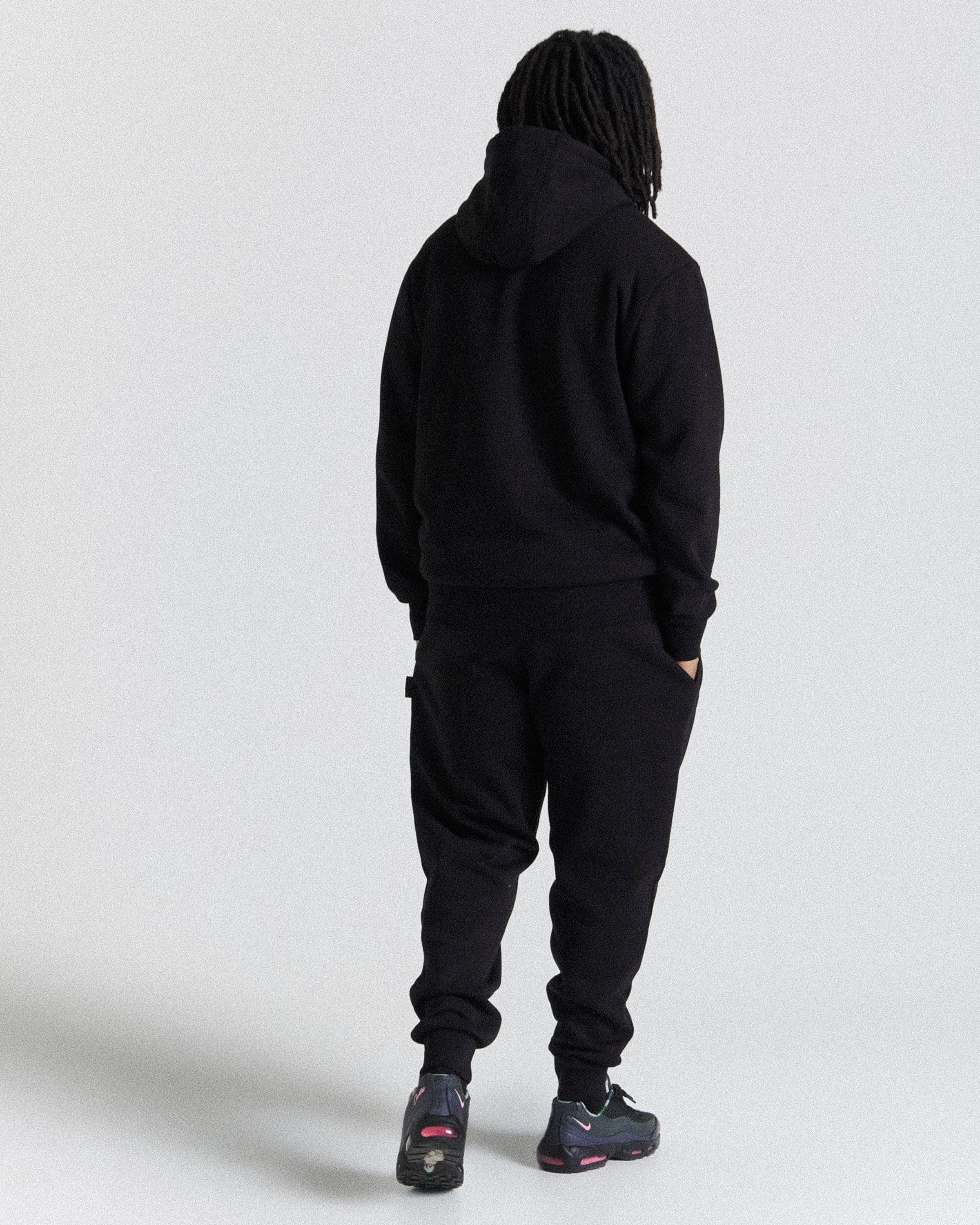 A person shown from the back wearing a black hooded sweatshirt and black jogger pants against a light gray studio background.