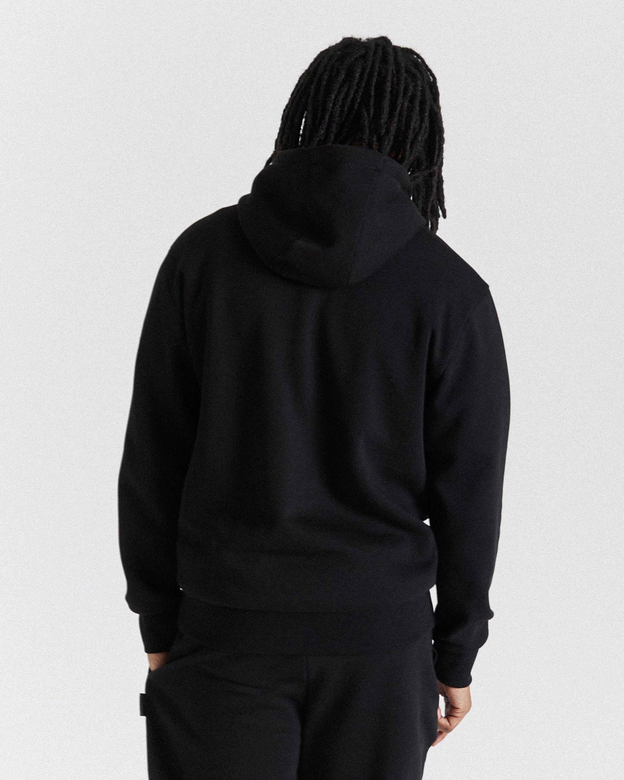 A person viewed from the back wearing a black pullover hoodie and black pants against a plain light gray studio background.