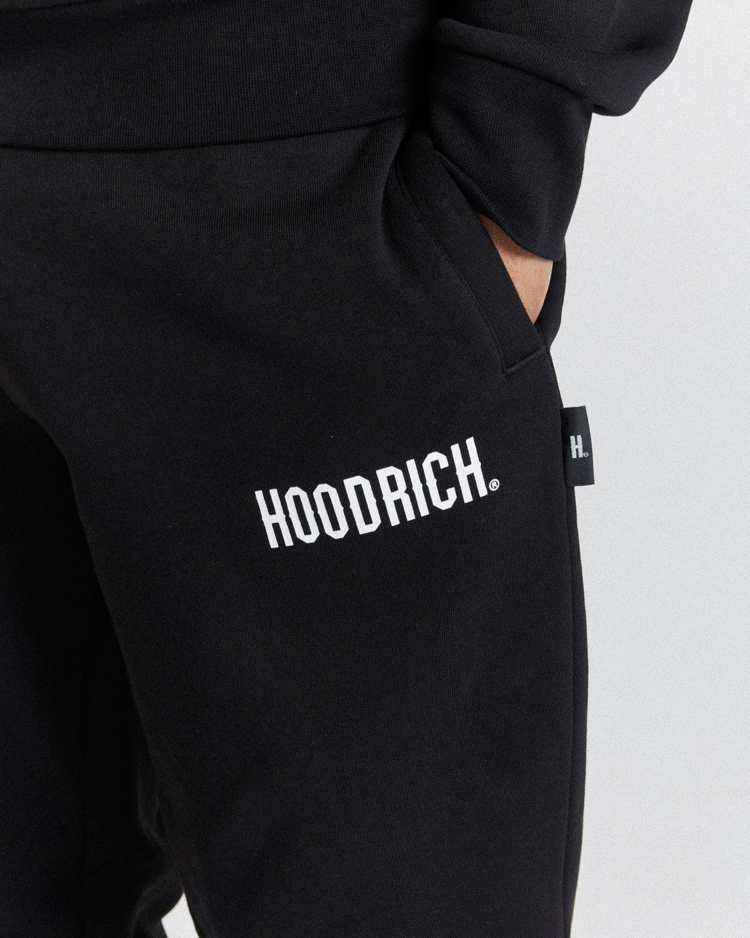 Close-up of black sweatpants with a white 'HOODRICH.' logo printed on the thigh and a small black side tag, shown against a light gray background.