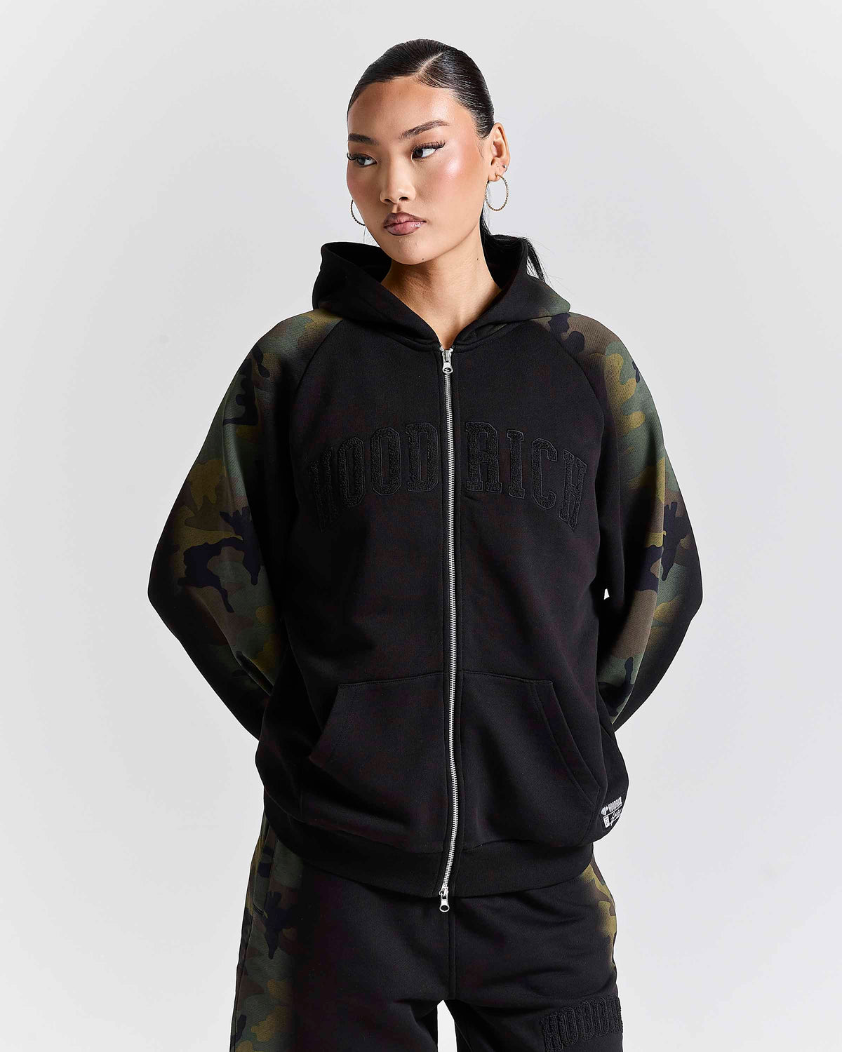 A model wearing a black full-zip hoodie with camouflage patterned raglan sleeves and a tonal black 'HOODRICH' logo across the chest, paired with matching camouflage pants, against a light gray studio background.