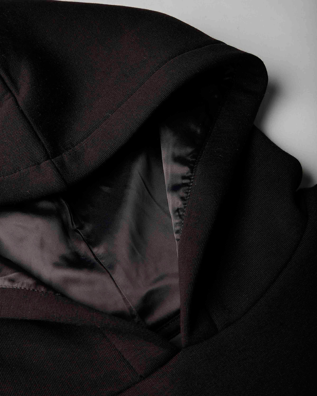 Close-up detail of a black hooded jacket or coat with a satin-like black interior lining, shown against a light gray background.