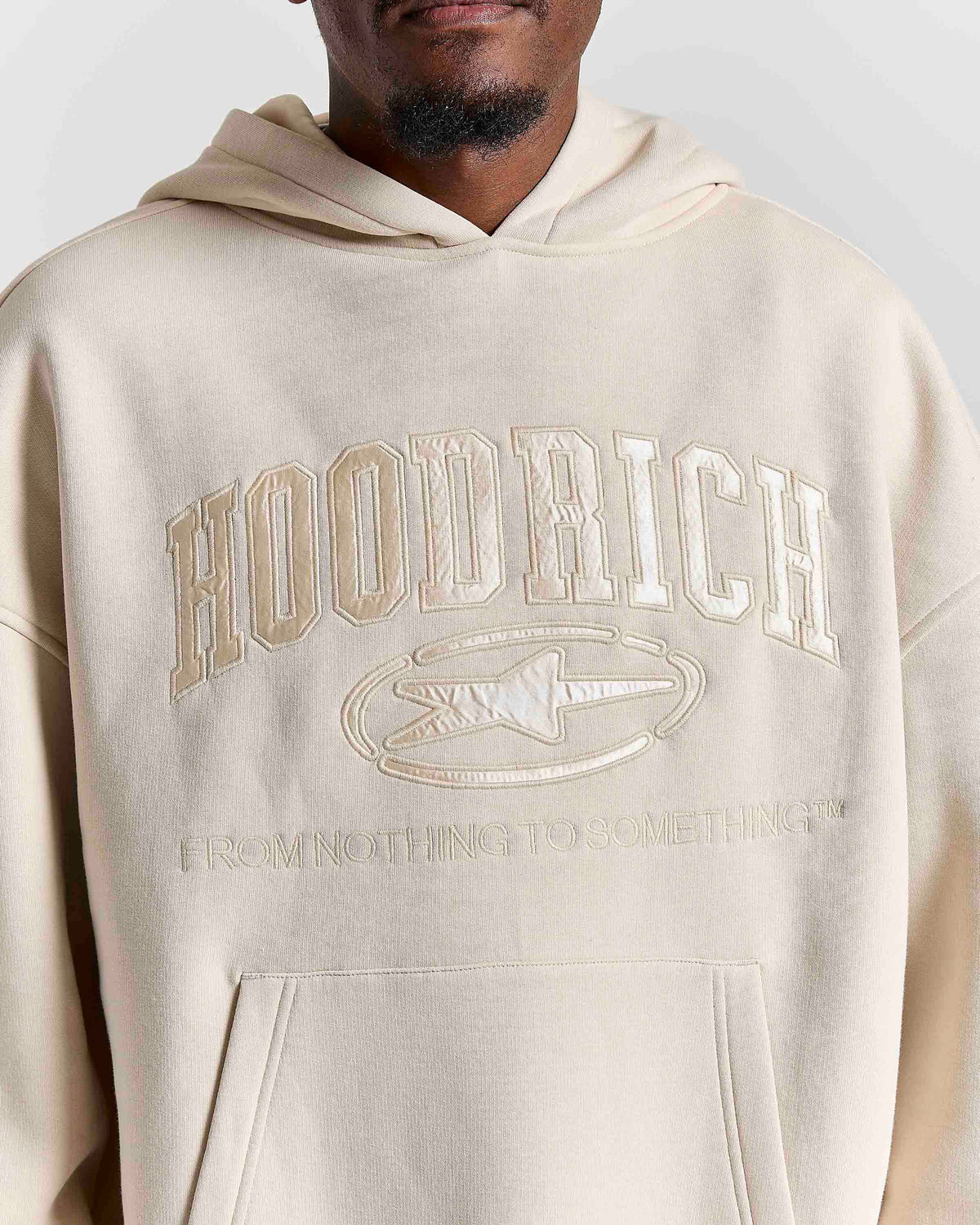 A close-up of a person wearing a cream-colored hoodie with tonal embroidered text reading 'HOODRICH' and 'FROM NOTHING TO SOMETHING' on the front.