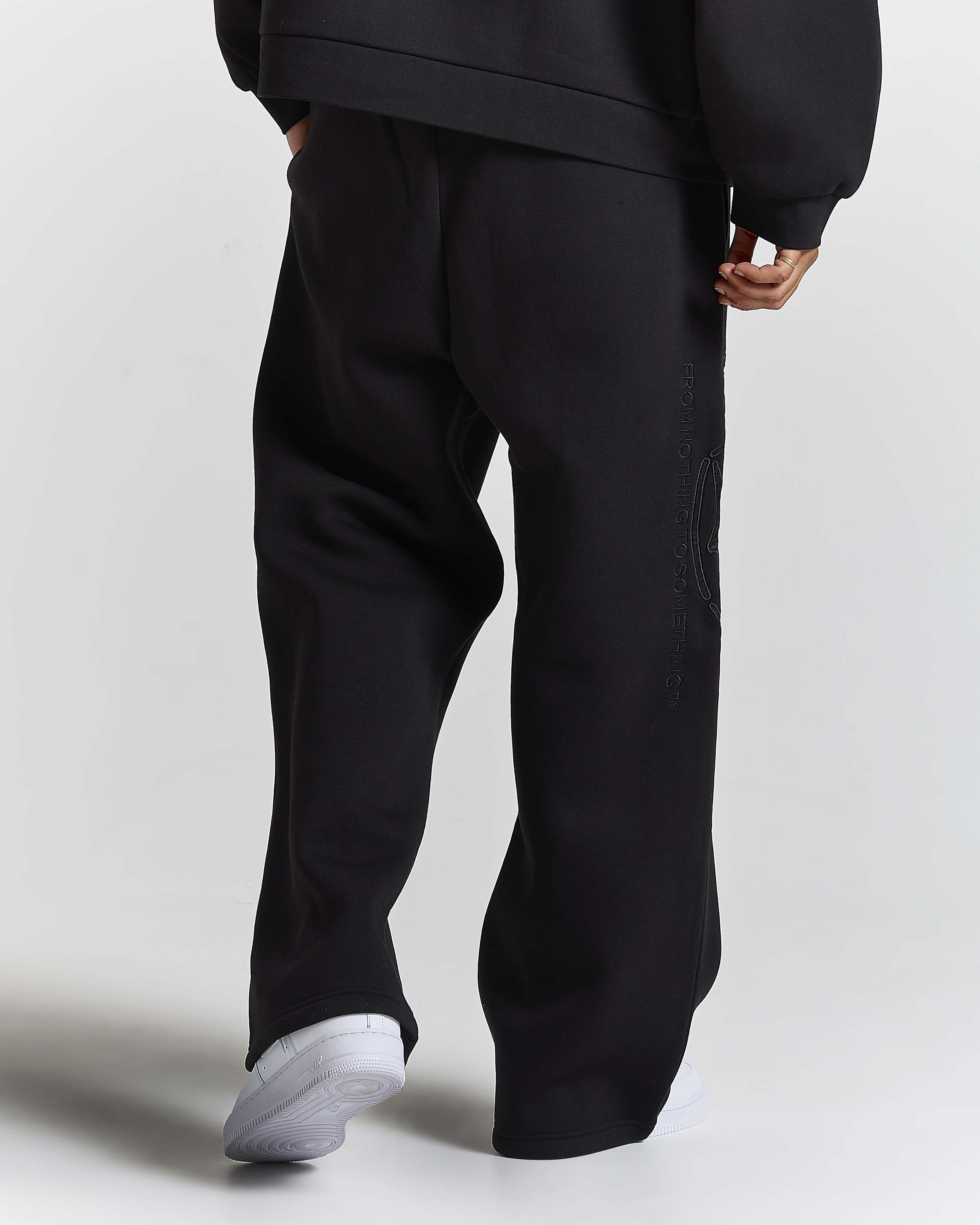 A person shown from the back wearing loose black sweatpants and a black sweatshirt, with white sneakers, against a light gray studio background.