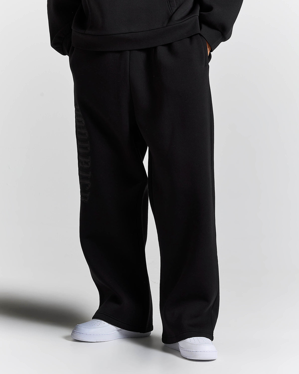 Black wide-leg sweatpants with a tonal vertical logo print on the left leg, shown on a model wearing white sneakers against a light gray background.