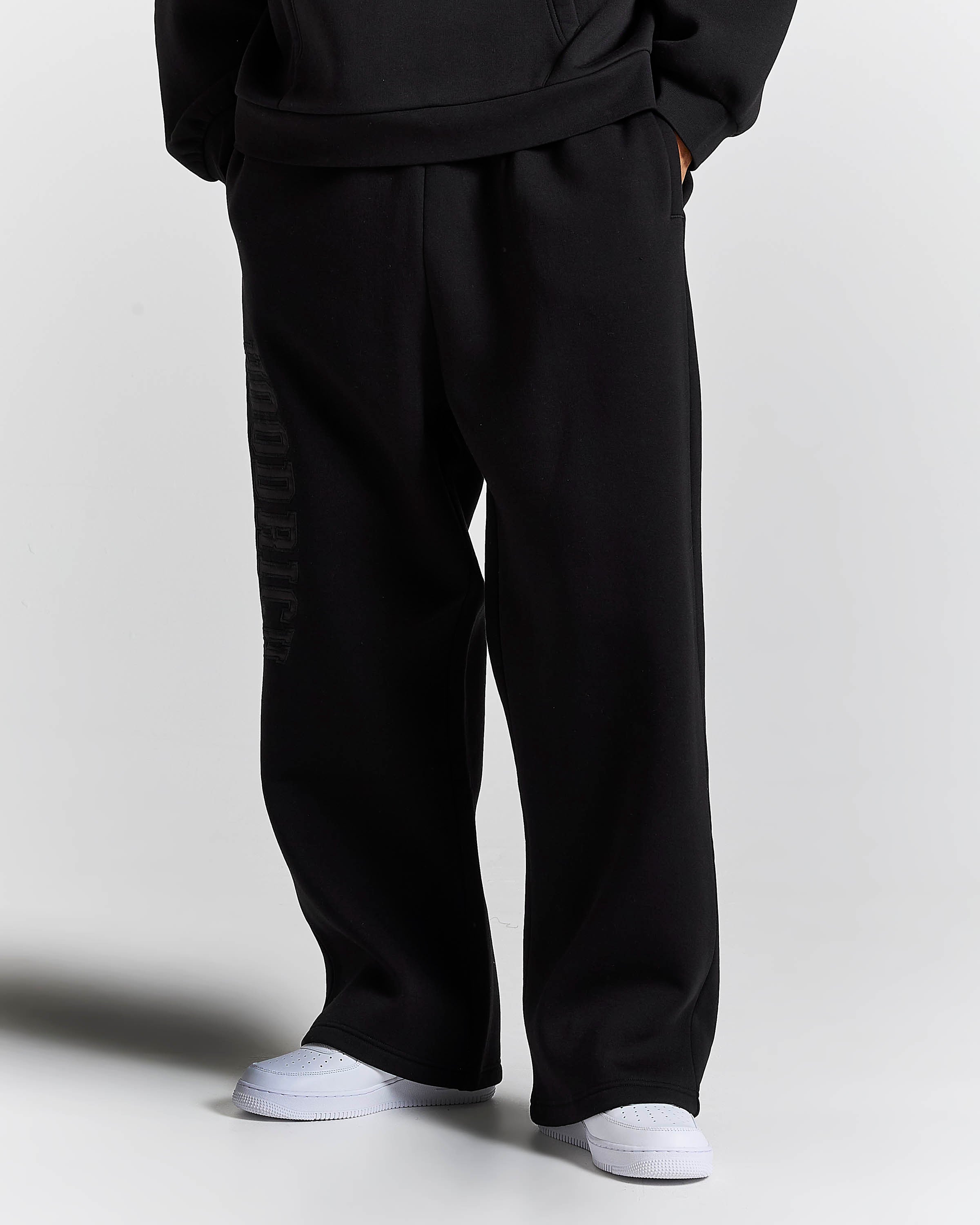 Black wide-leg sweatpants with a tonal vertical logo print on the left leg, shown on a model wearing white sneakers against a light gray background.