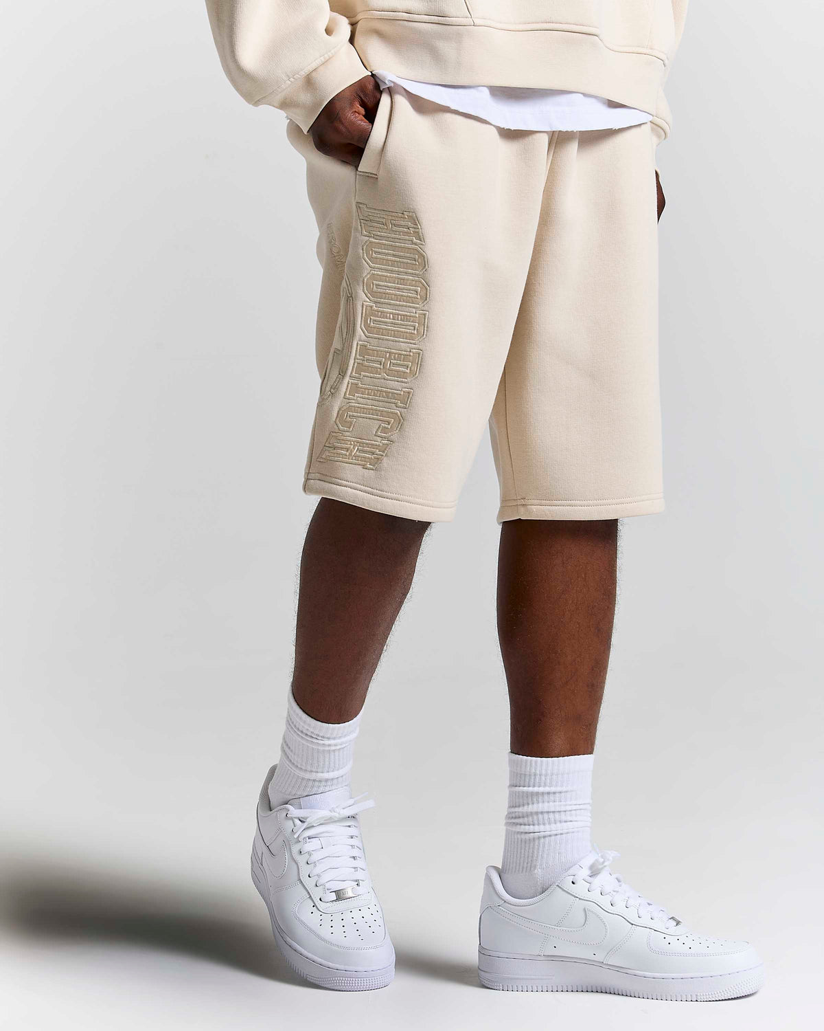 A person wearing beige sweat shorts with a metallic gold embroidered HOODRICH logo down the side, styled with a white t-shirt, white crew socks, and white Nike Air Force 1 sneakers against a light gray studio background.