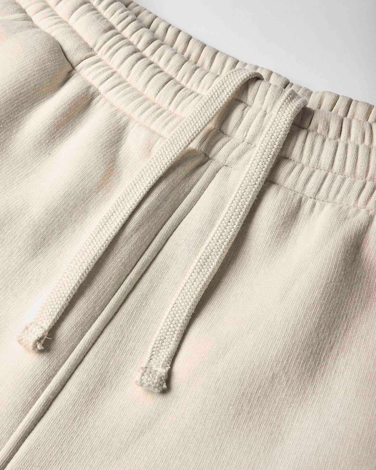 Close-up of a pair of light beige or cream sweatpants showing the textured fabric, elastic waistband, and matching drawstrings.