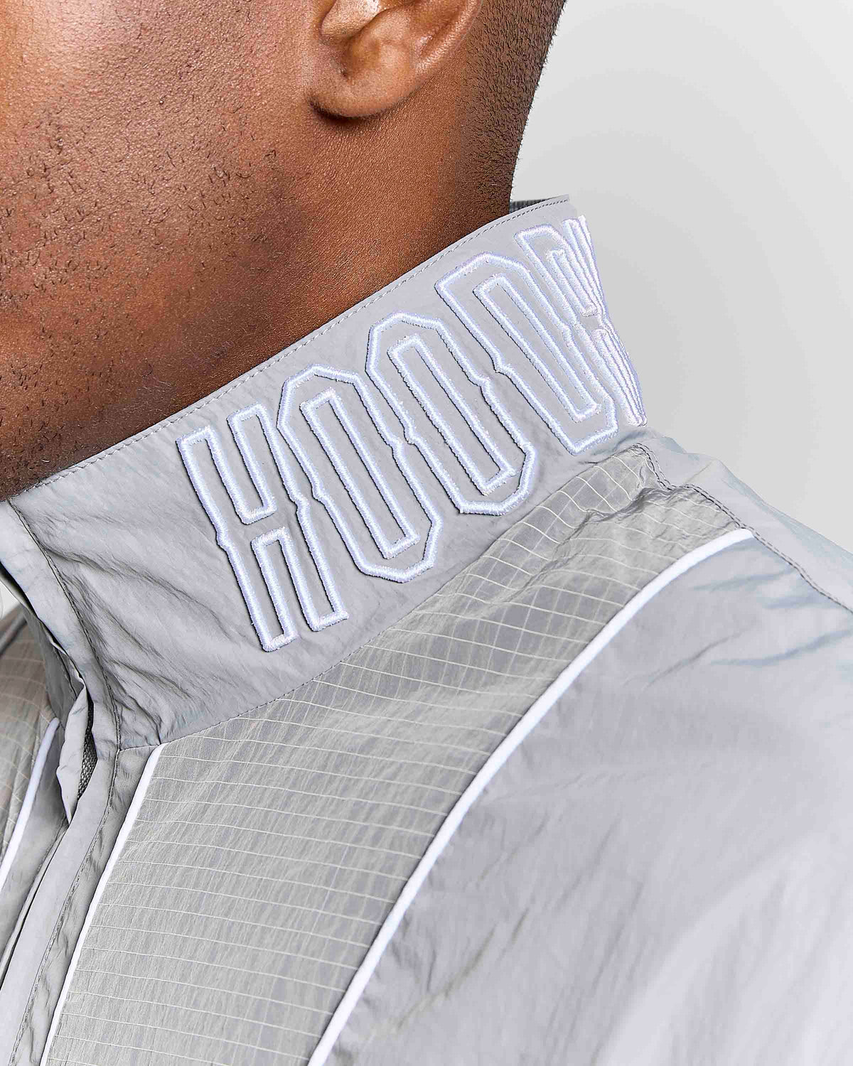 Close-up of a model wearing a light gray hooded windbreaker jacket, highlighting the high collar with white embroidered 'HOODRICH' lettering.