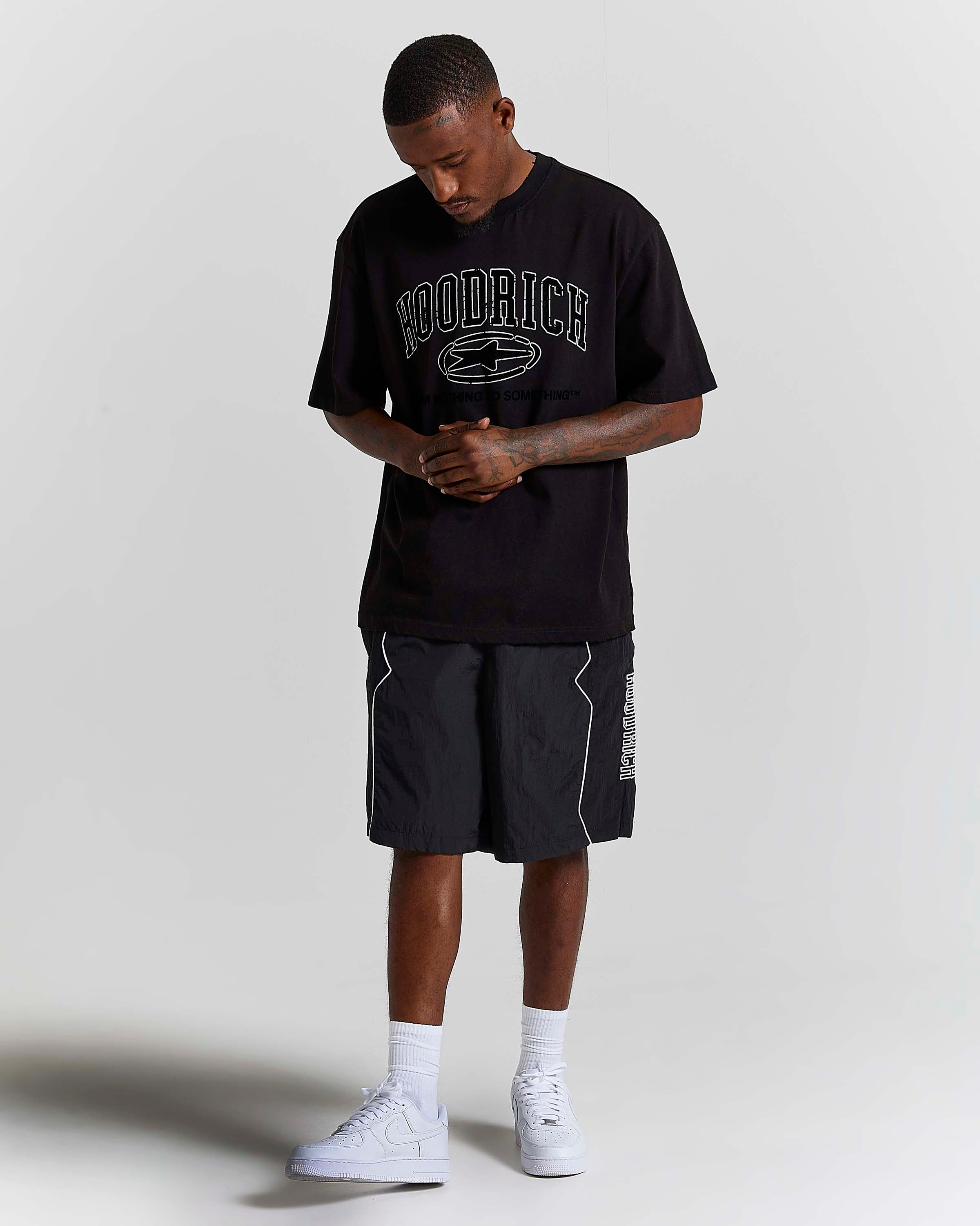 A man modeling a black Hoodrich t-shirt with a white graphic logo, paired with black athletic shorts, white socks, and white sneakers, standing against a light gray studio background.