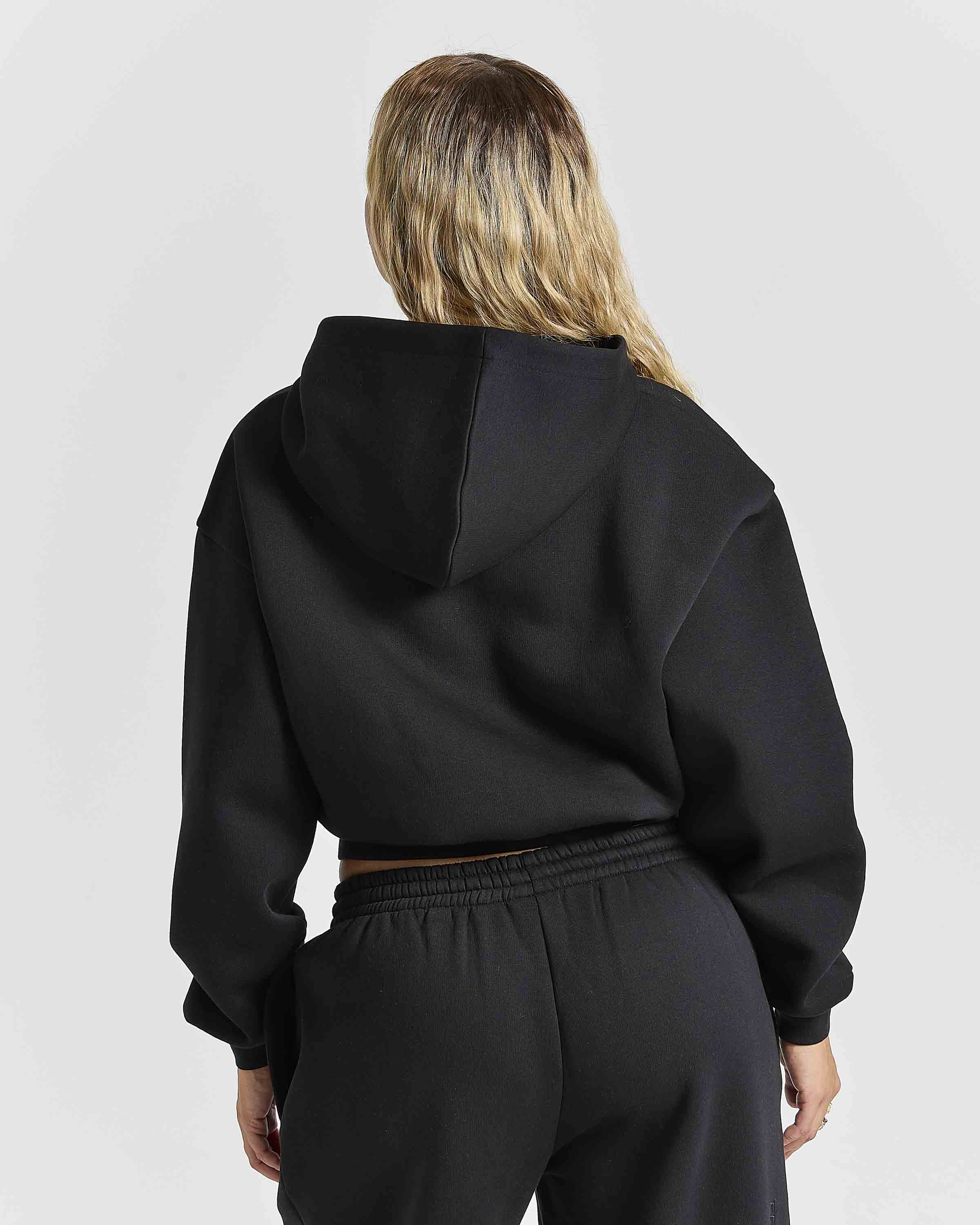 A woman viewed from behind wearing a black cropped hoodie and black sweatpants against a light gray studio background.