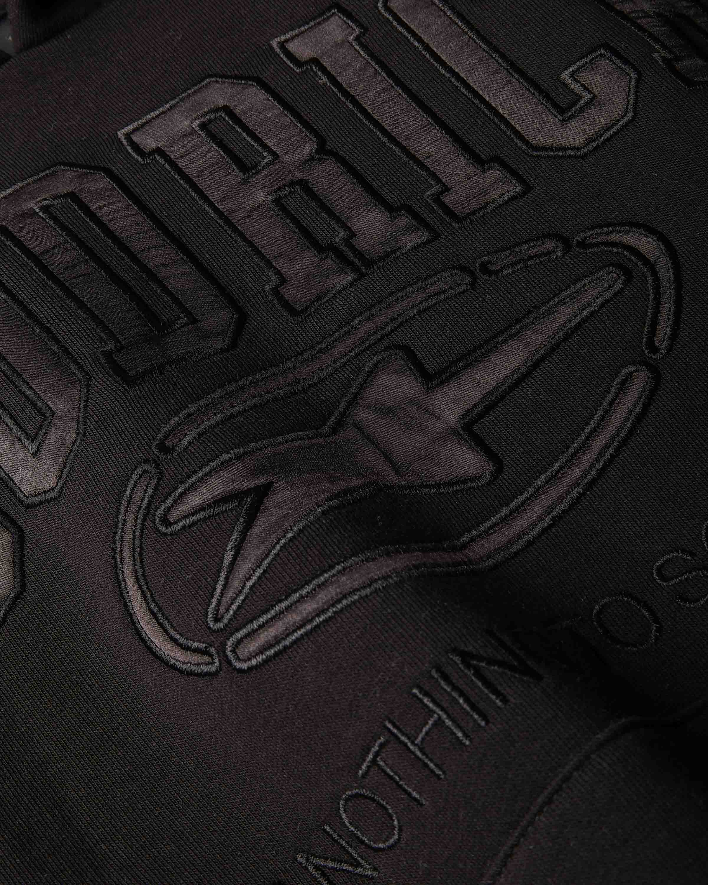 Close-up of a dark embroidered graphic on fabric, showing large raised letters and a stylized logo in a very dark brown or charcoal tone against a black background.