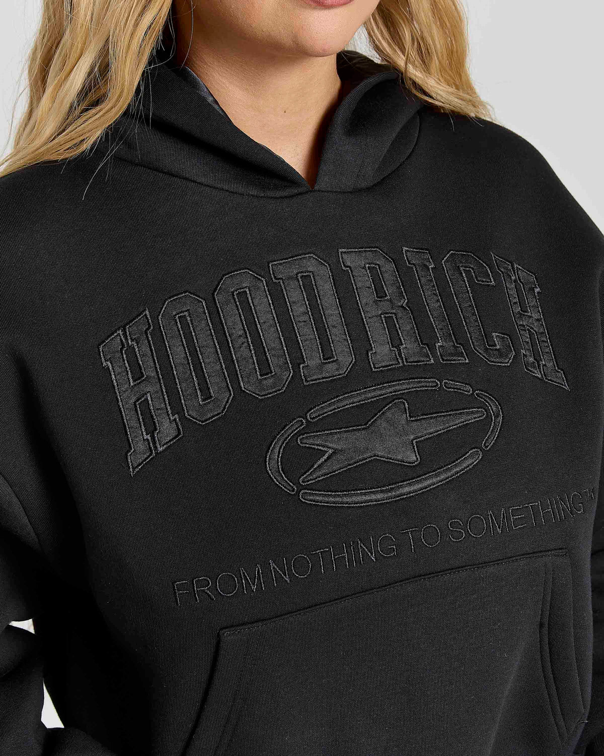Close-up of a person wearing a black hoodie with tonal dark gray 'HOODRICH' lettering and a star logo on the chest, plus smaller text reading 'FROM NOTHING TO SOMETHING'.