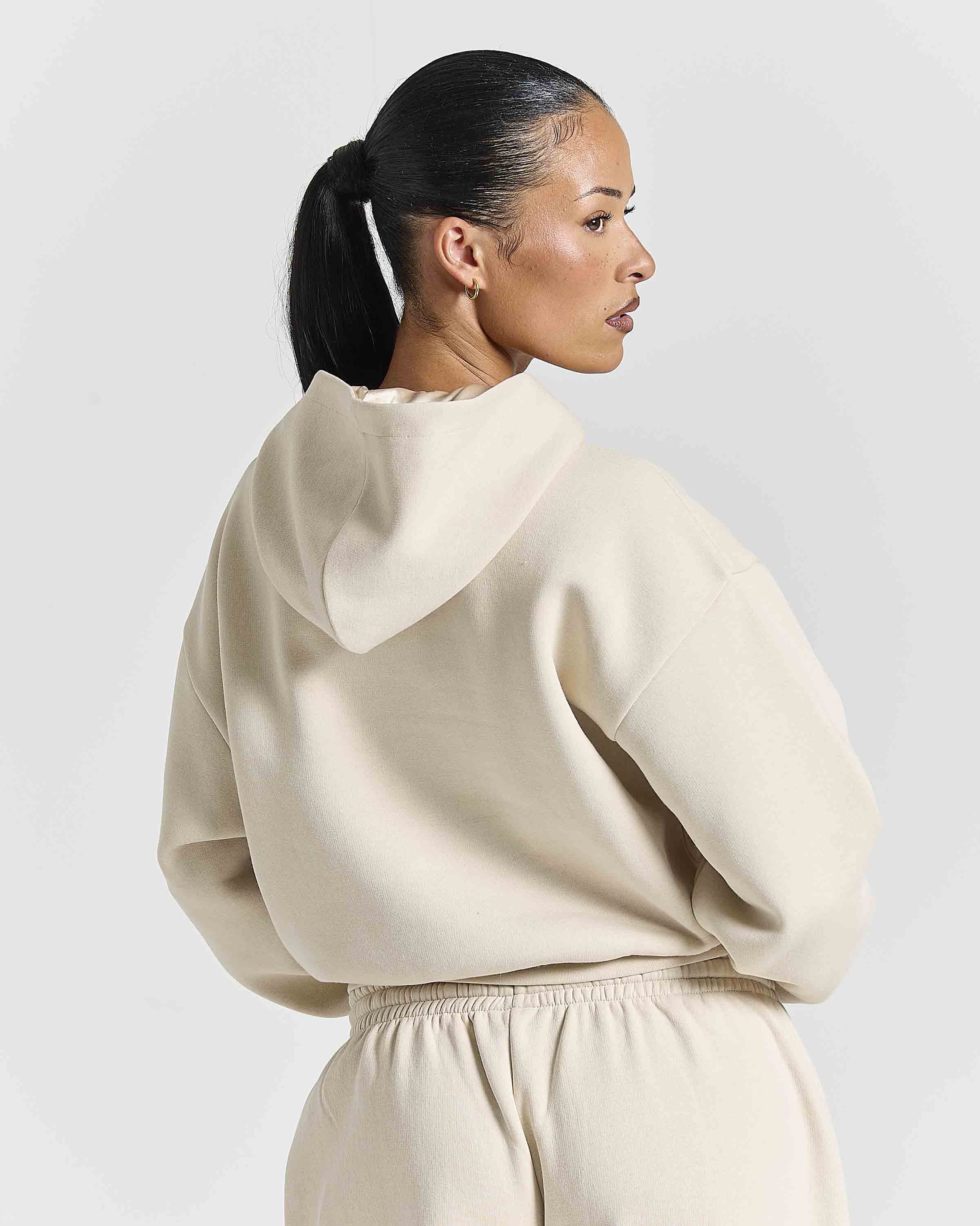 A woman with her hair in a ponytail is shown from the back wearing a cream-colored oversized hoodie and matching sweatpants against a light gray studio background.