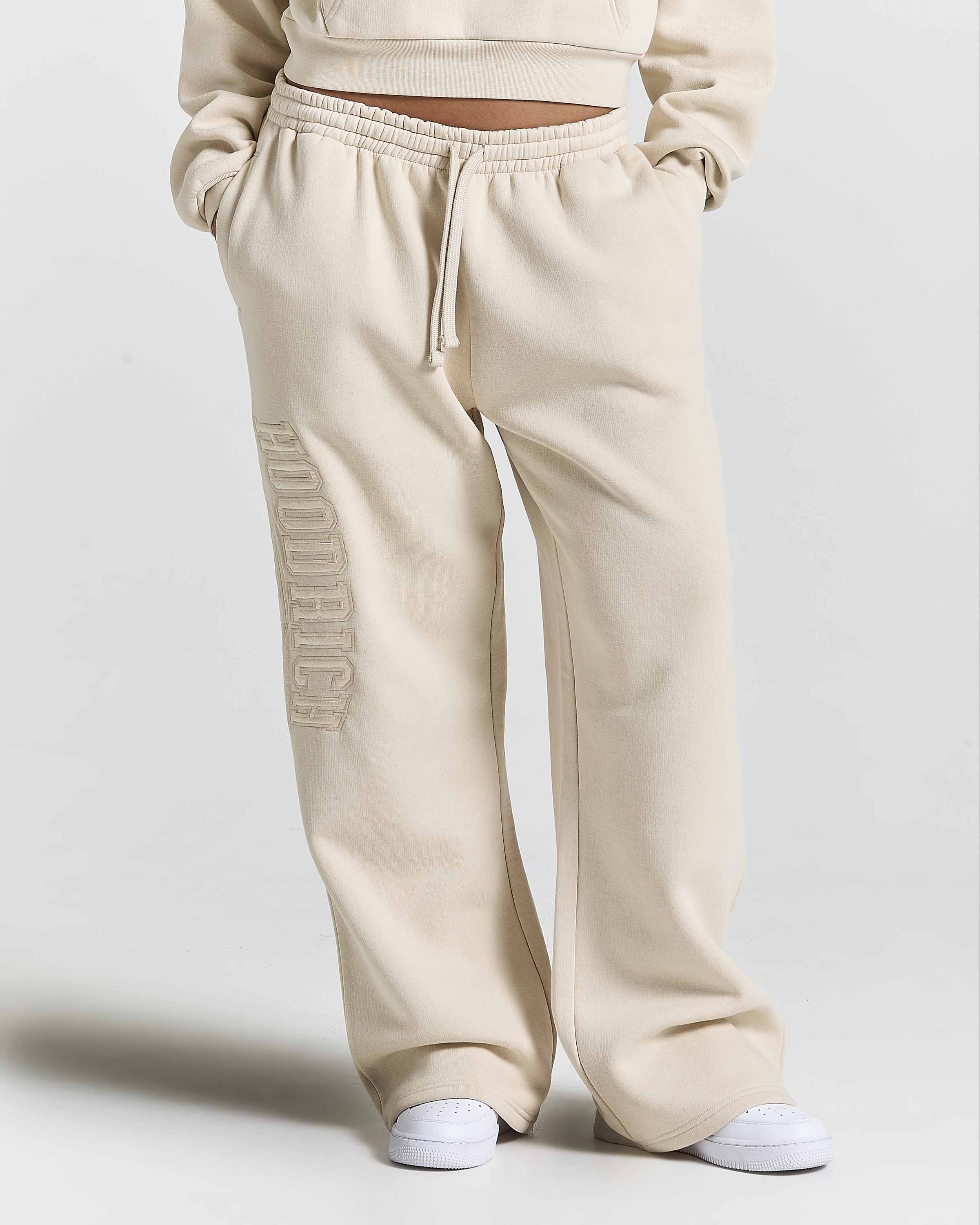 A person wearing cream-colored wide-leg sweatpants with a vertical tonal 'HOODRICH' logo on the right leg, paired with white sneakers against a light gray studio background.