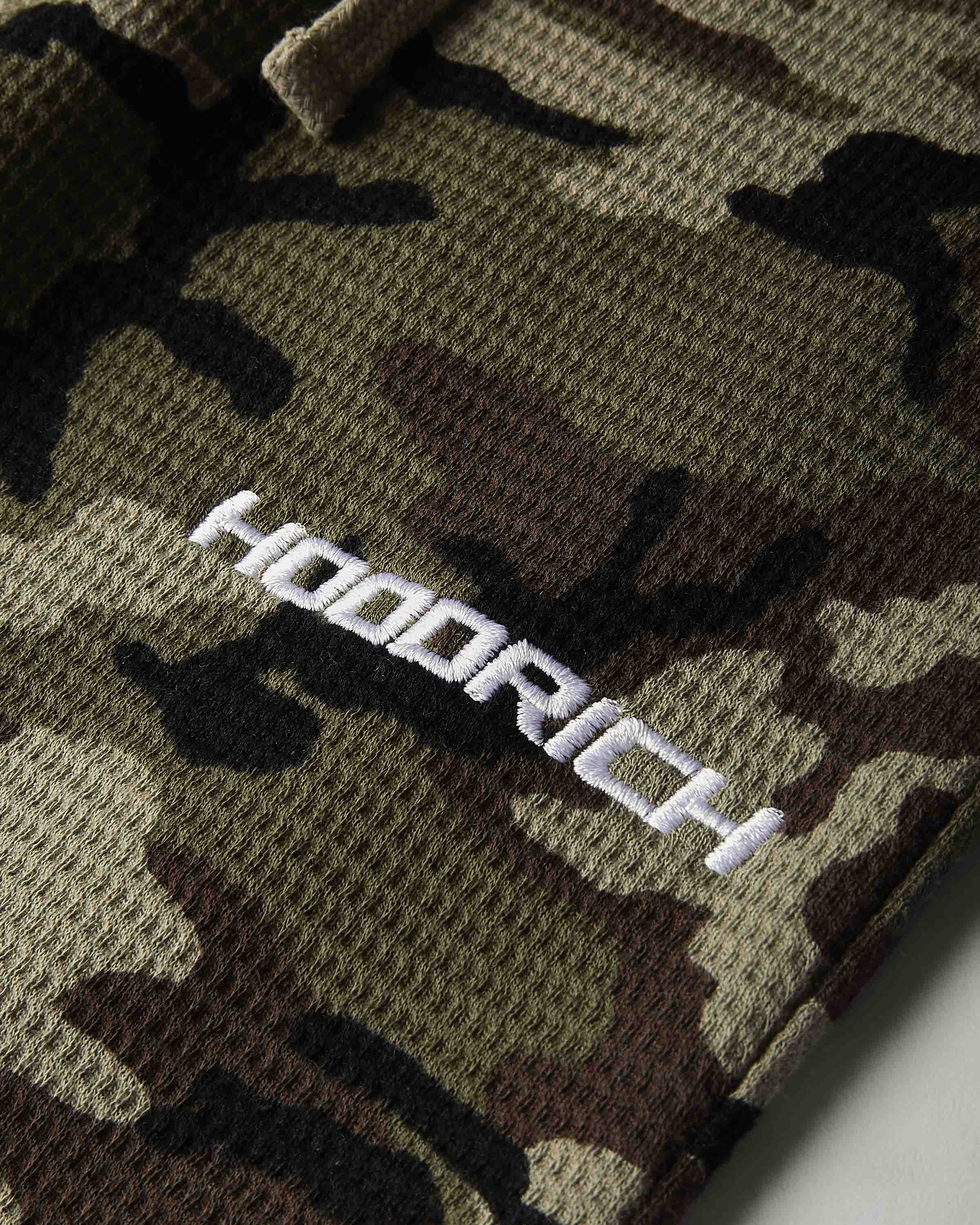 Close-up of a camouflage knit garment with a white embroidered 'HOODRICH' logo on the chest.