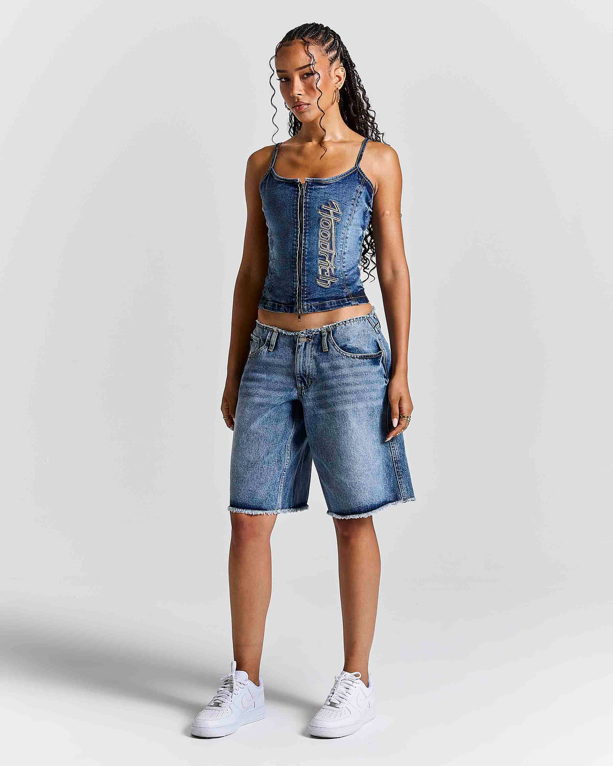 A woman modeling a matching denim set consisting of a blue denim camisole top with silver embellishments and blue denim shorts, paired with white sneakers, against a light gray studio background.