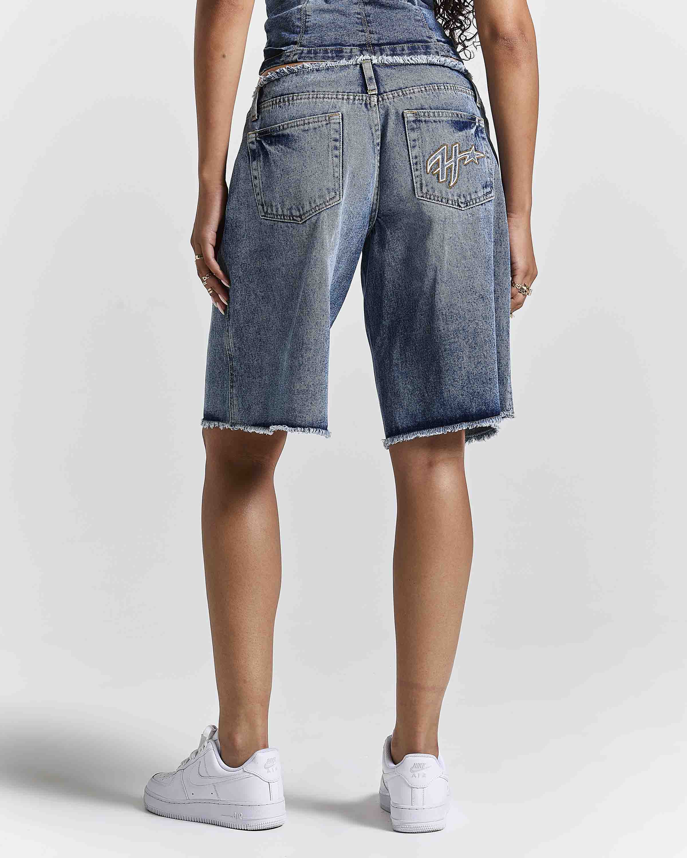 A person shown from the back wearing medium-wash blue denim shorts with a raw frayed hem, paired with white sneakers against a light gray studio background.