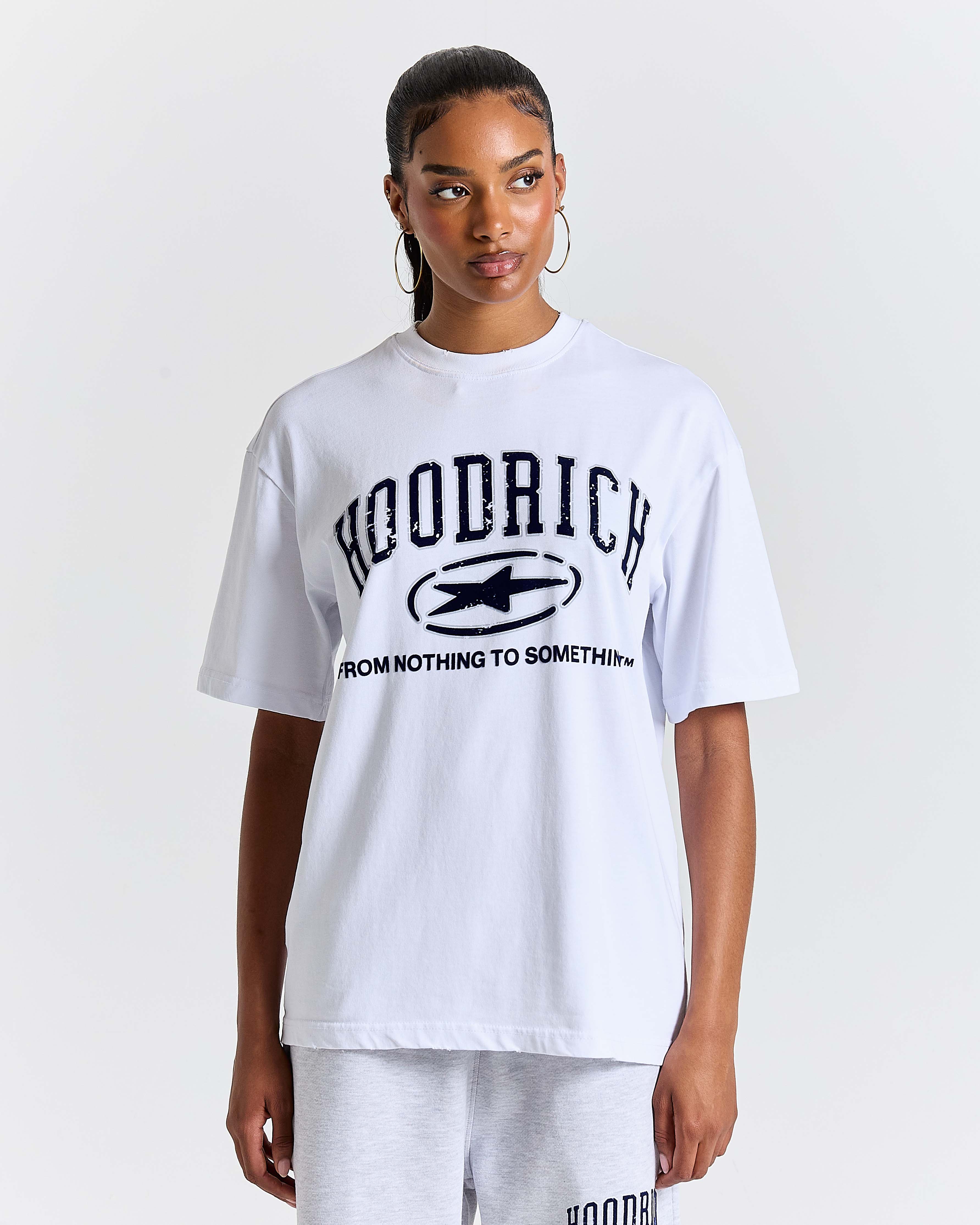 Female model wearing a white 'HOODRICH' t-shirt with a star logo and 'FROM NOTHING TO SOMETHING' spell-out on a plain background