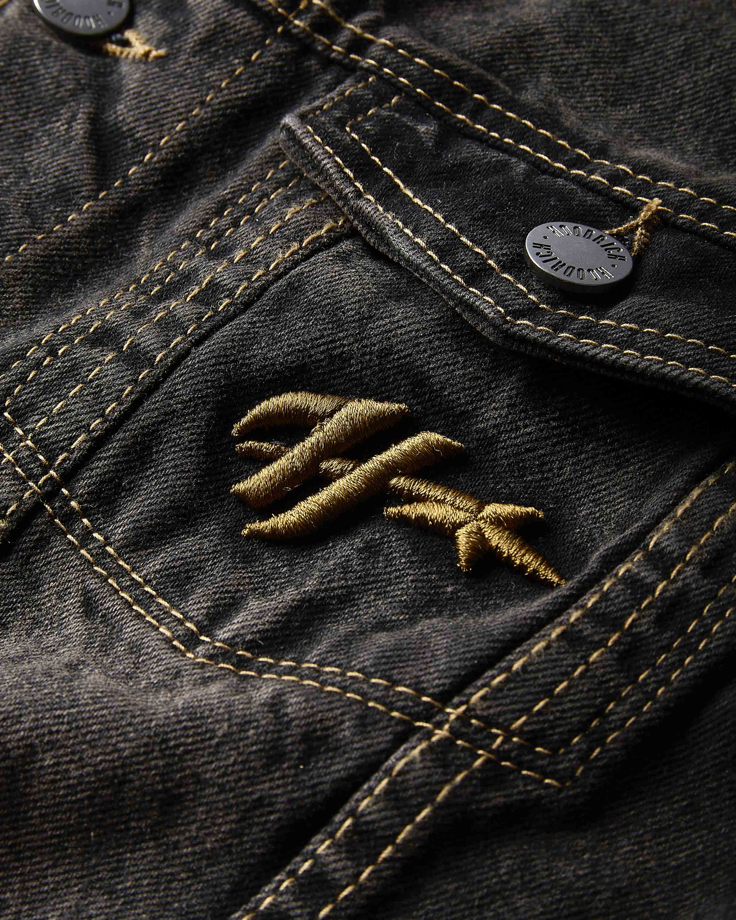 Close-up of the Hoodrich logo embroidered on the chest pocket of the black rust washed denim jacket.