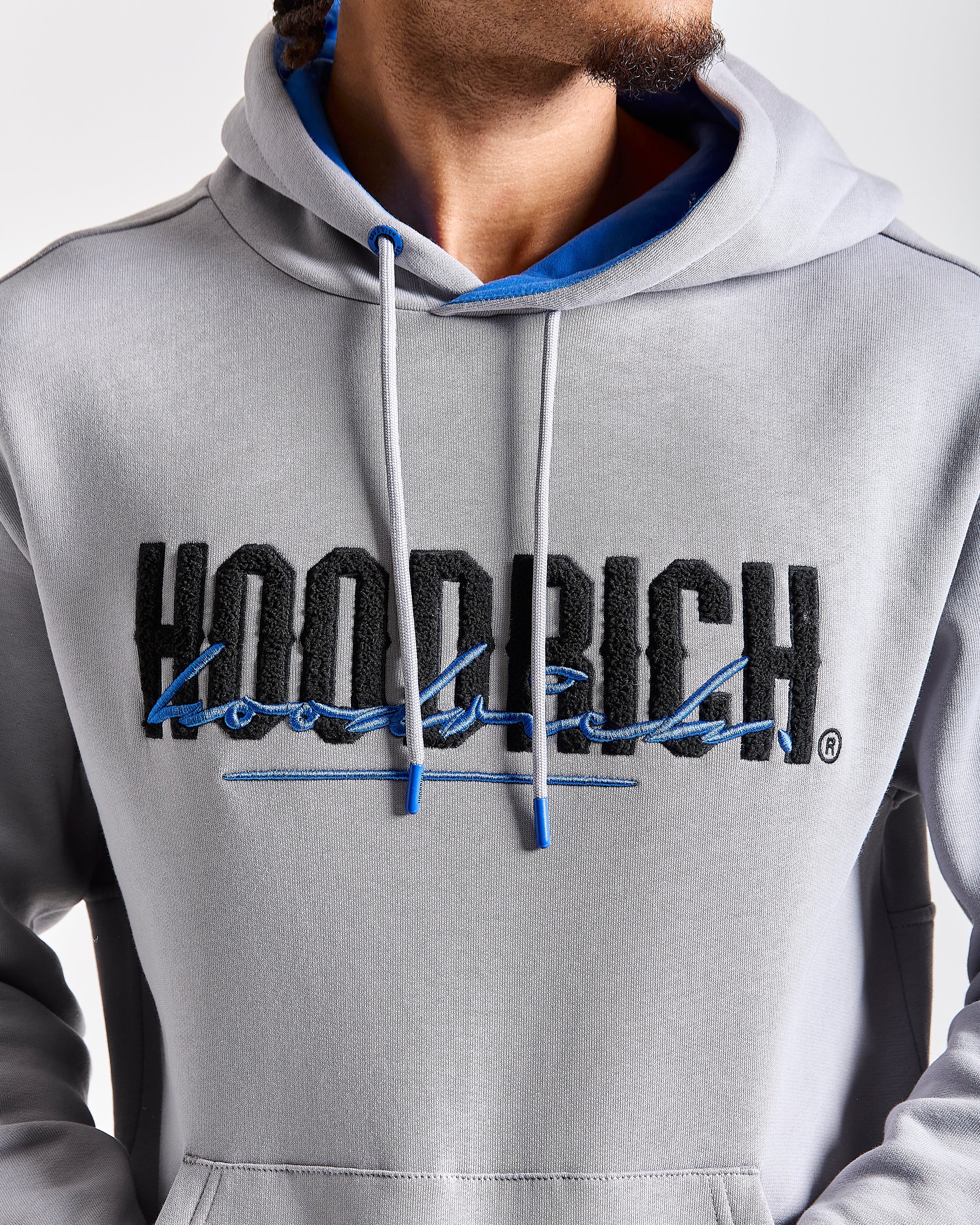 A close-up of a person wearing a light gray hoodie with a large black and blue 'HOODRICH' logo across the chest, featuring white drawstrings with blue tips.