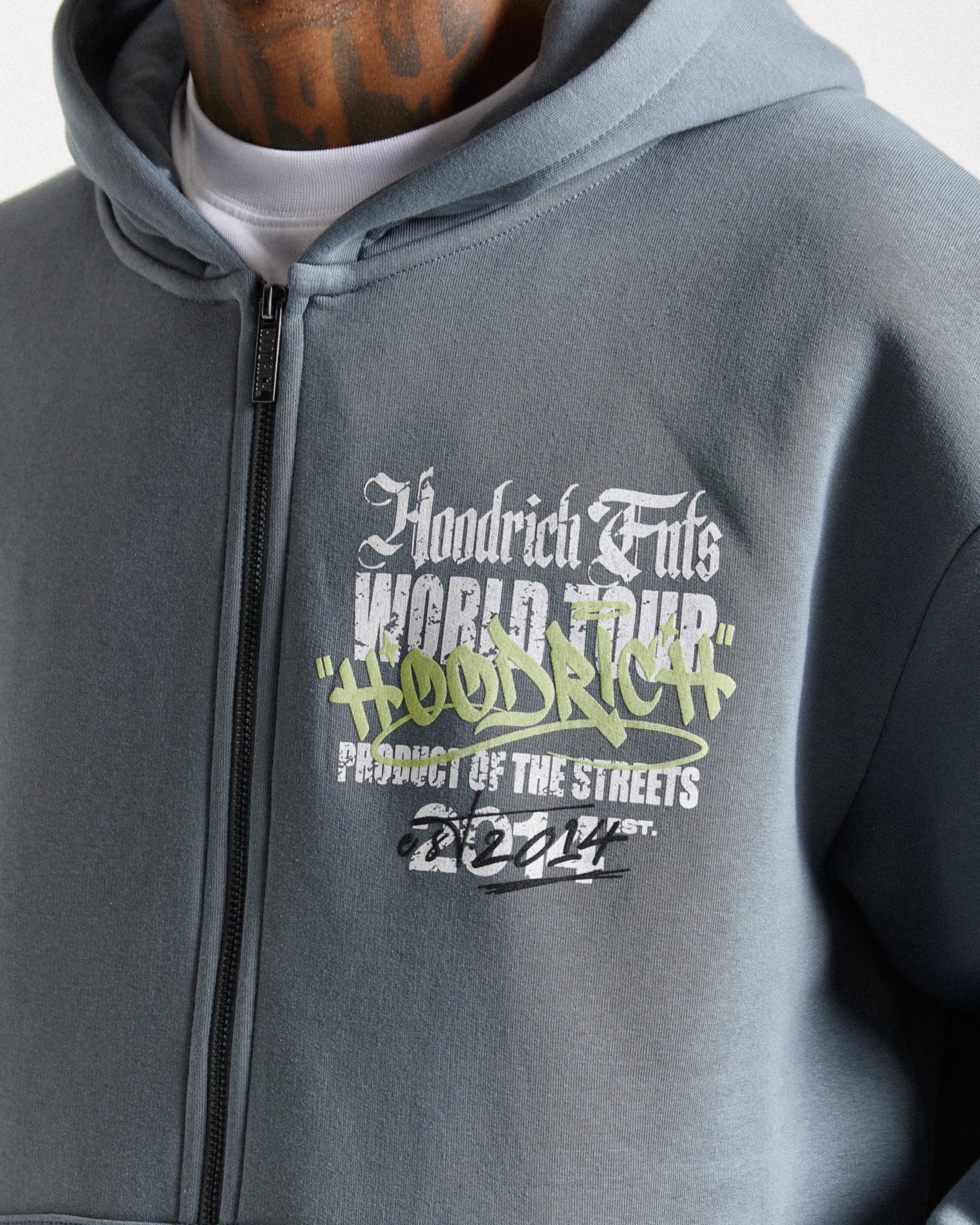 A close-up of a person wearing a heather gray zip-up hoodie with a graphic print on the chest. The graphic features the text 'Hoodrich Fils World Tour' in white and green, with 'Product of the Streets' and '2024' below.