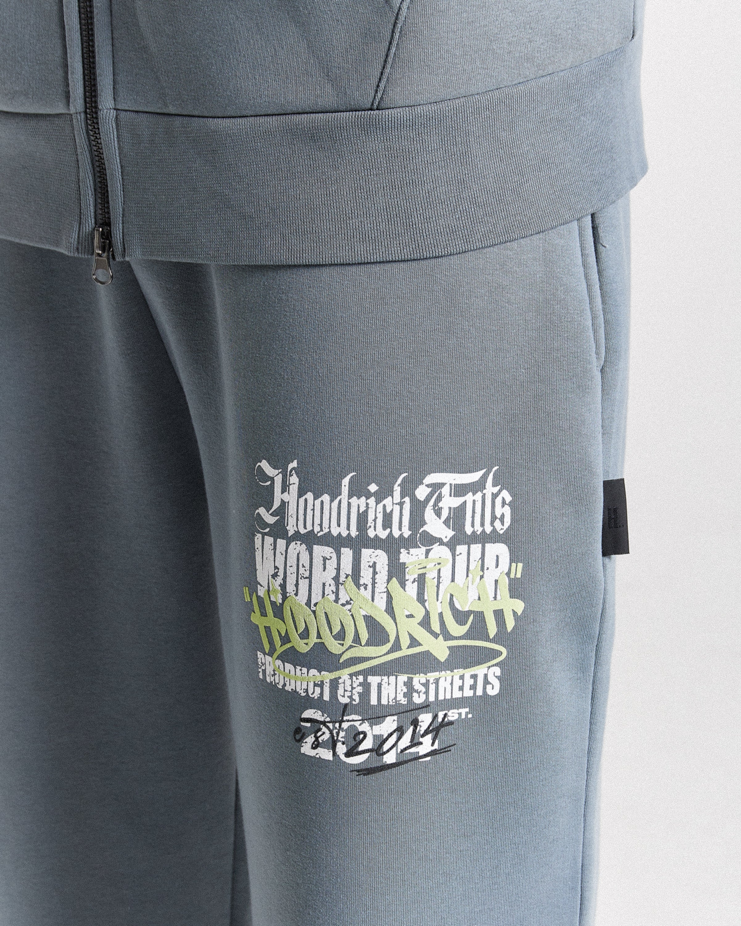 Close-up of a pair of heather gray sweatpants featuring a graphic print on the thigh. The graphic reads 'Floodrich Fuls World Tour' in white and lime green, with 'Product of the Streets' and '2024' below.