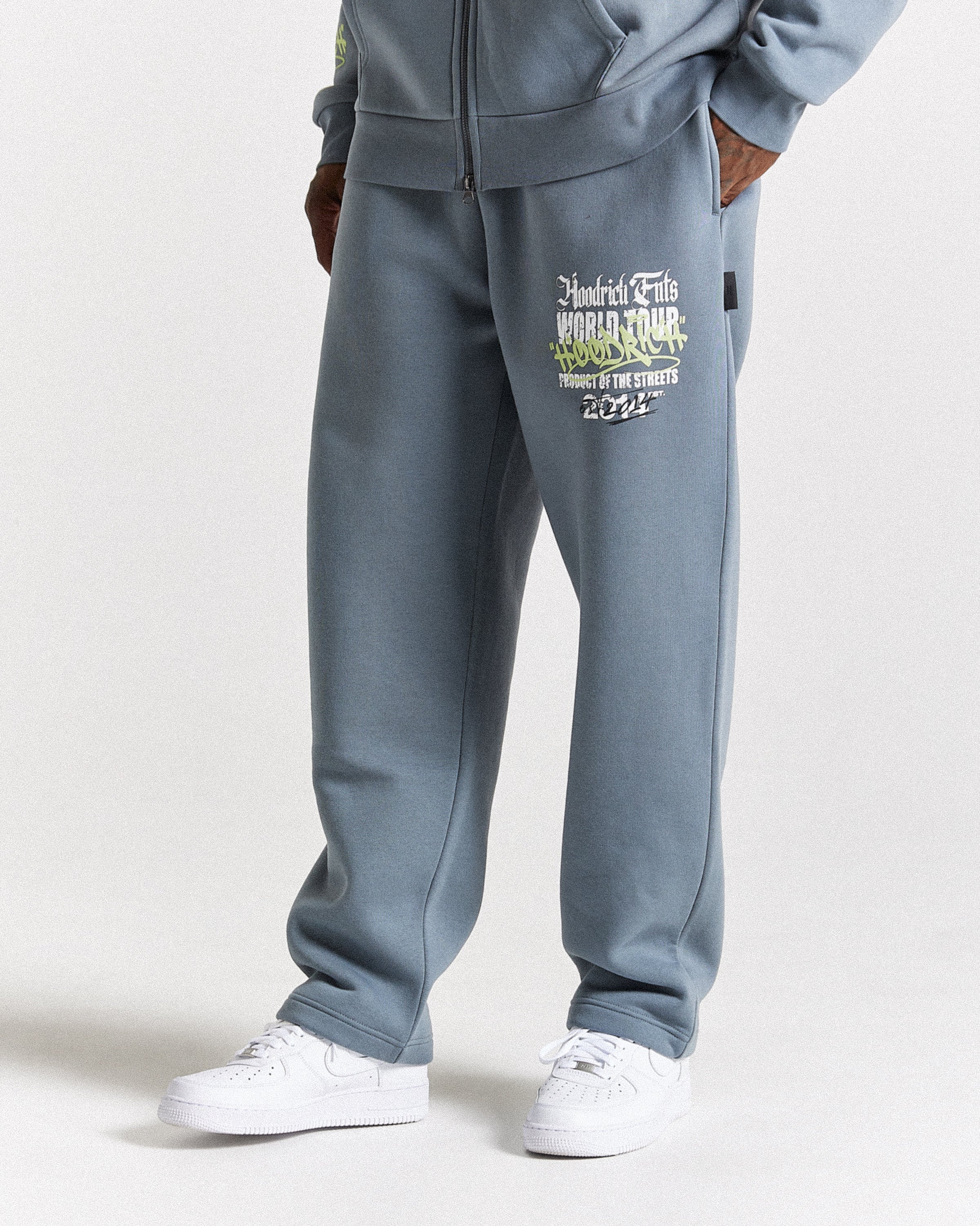 A person wearing slate blue sweatpants with a graphic print on the left thigh, paired with white sneakers, shown from the waist down against a light gray studio background.