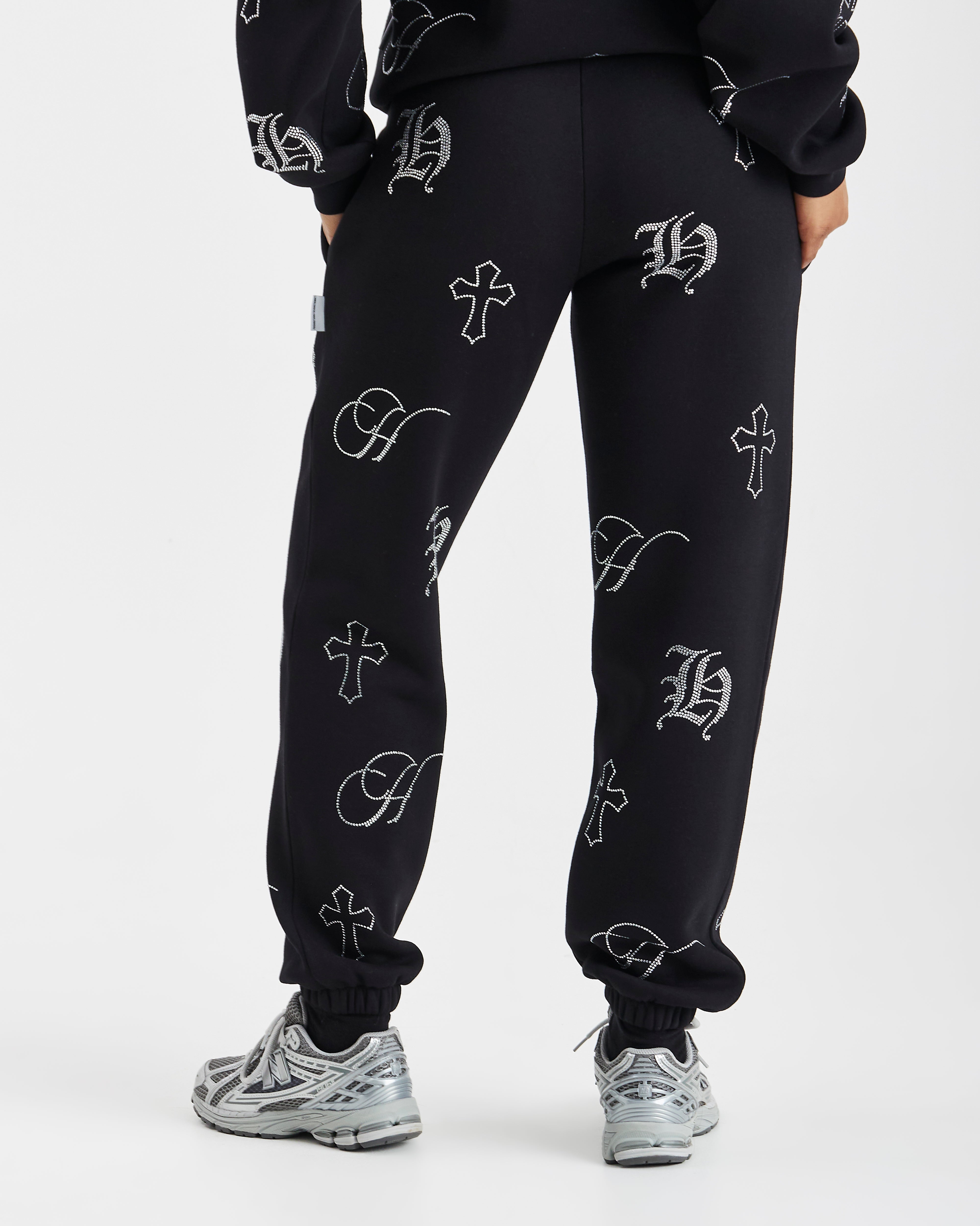 Black sweatpants with an all-over white Gothic monogram and cross pattern, shown on a model from the waist down wearing silver sneakers against a light gray background.