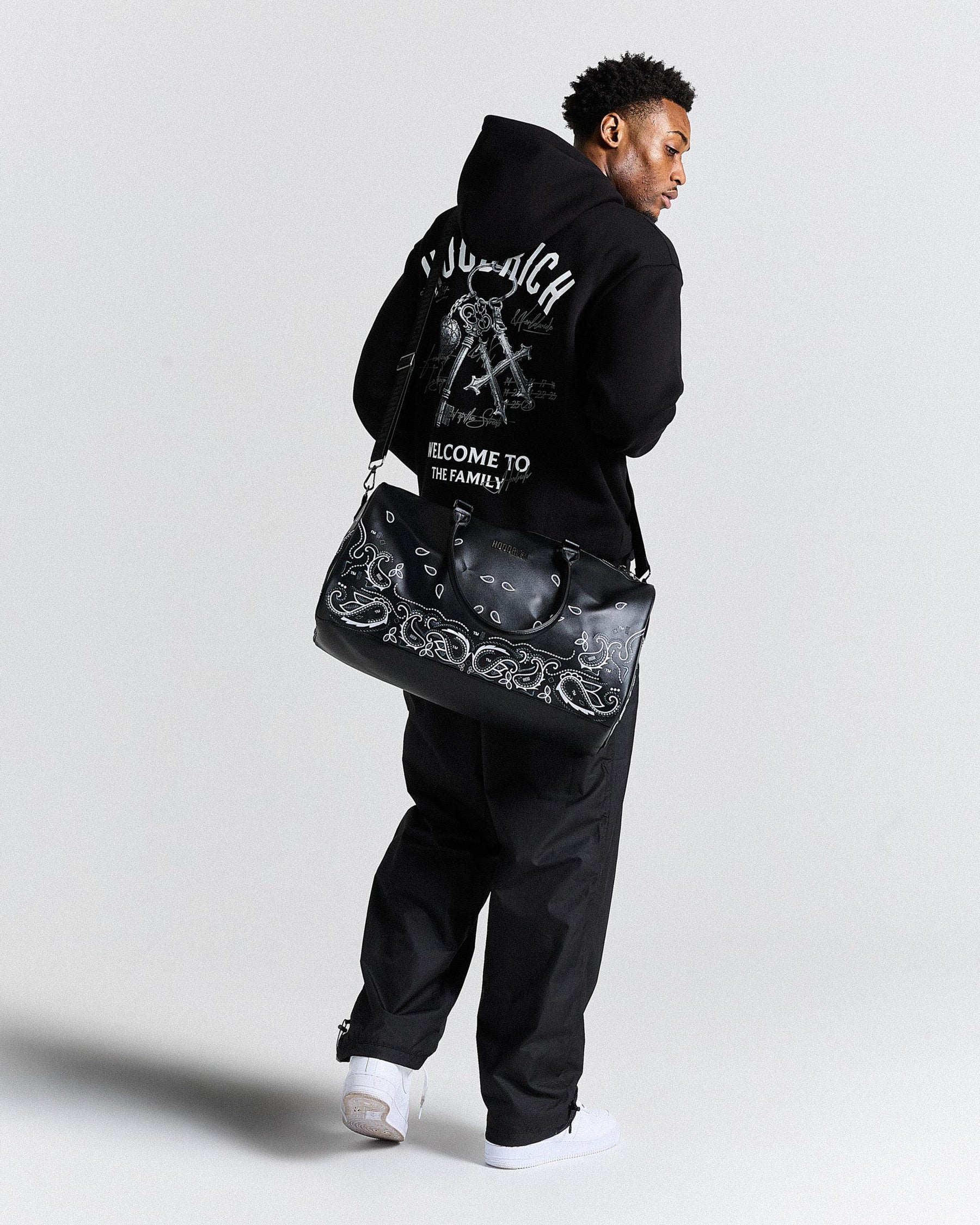 Person wearing a black hoodie and pants, holding a black bag with white designs on a light gray background