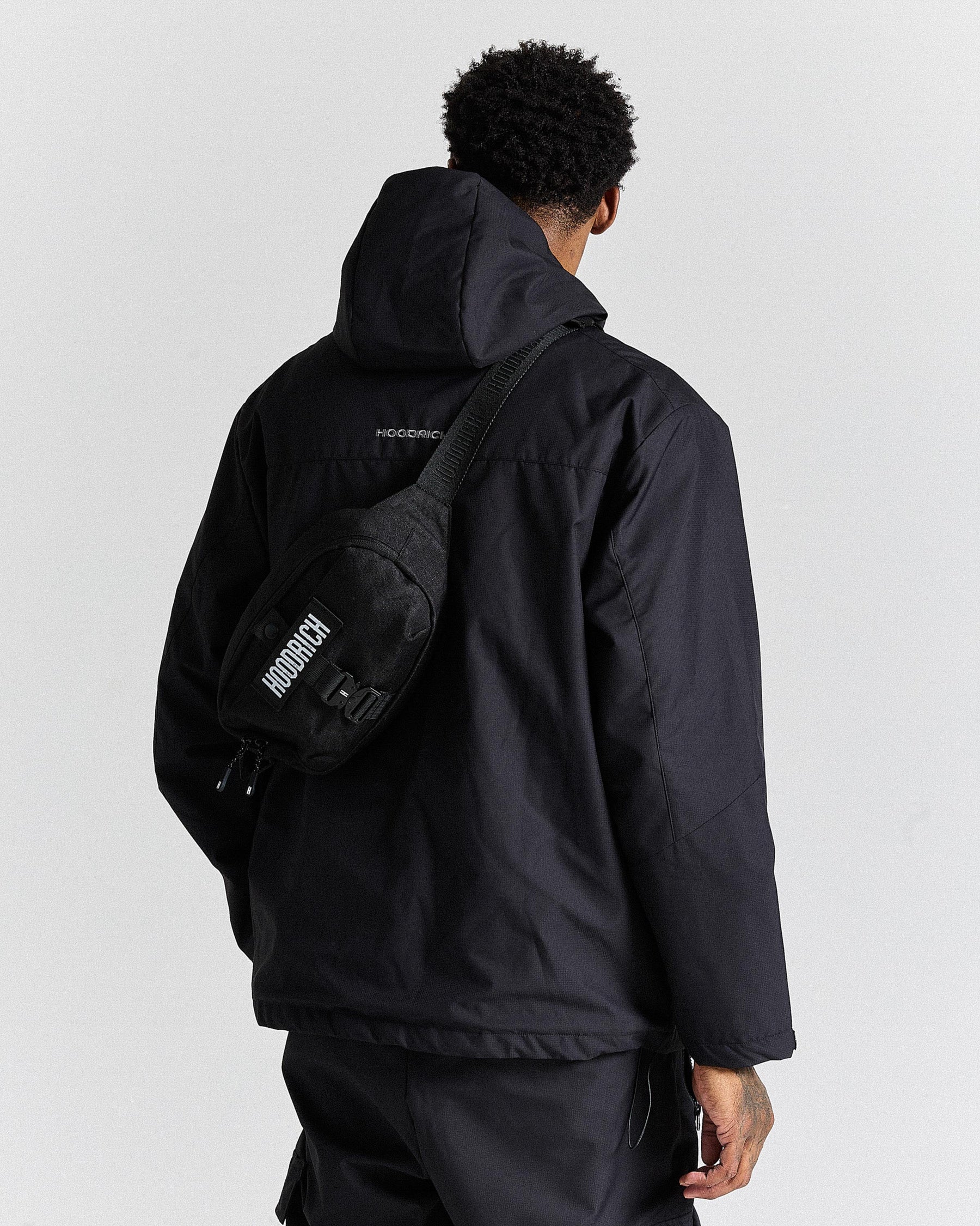 Person wearing a black jacket and carrying a black bag with 'Hoodrich' branding on a light gray background